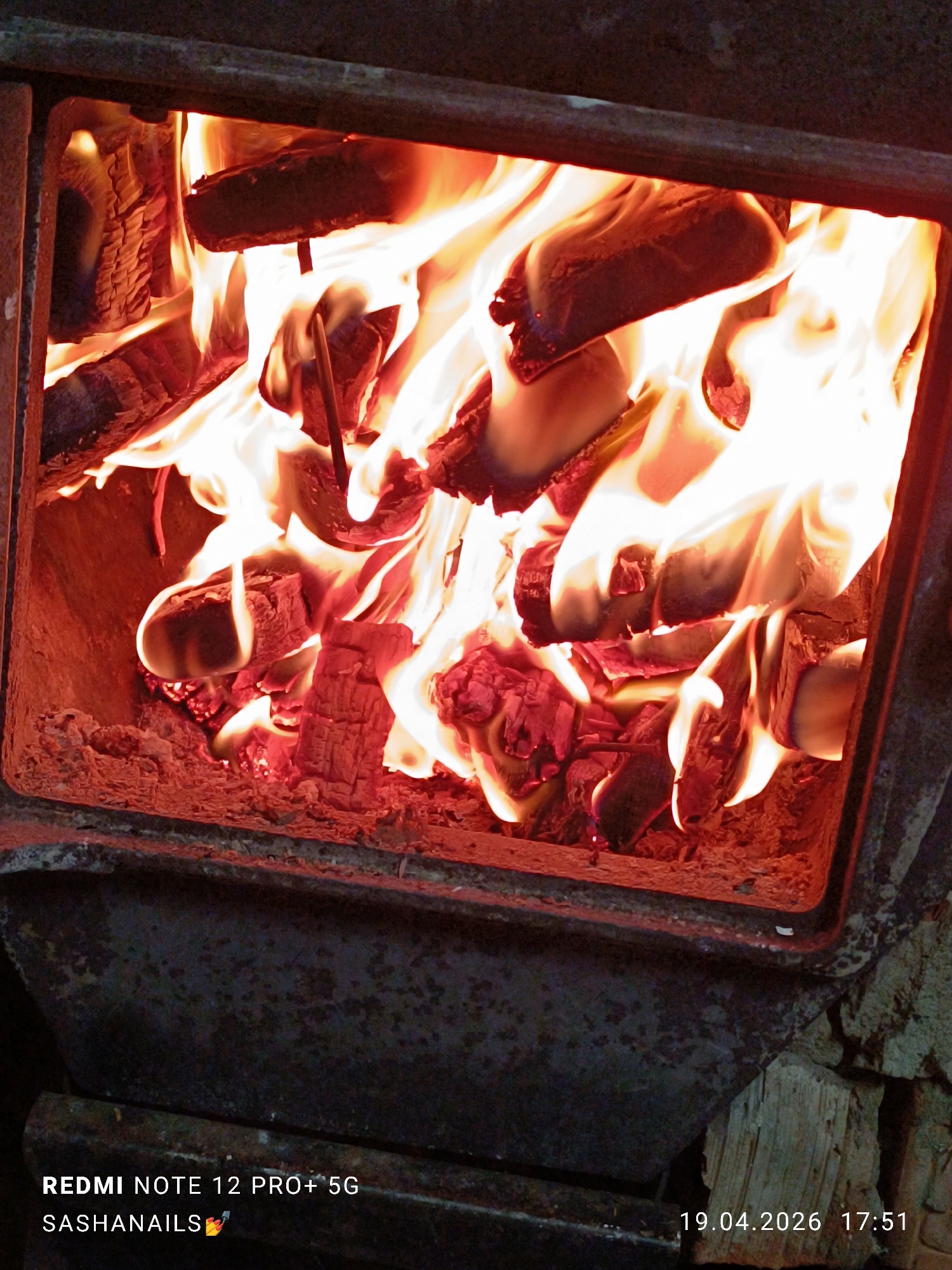 The image displays a close-up view of a roaring fire contained within a metal fireplace or wood-burning stove. Bright, undulating flames in shades of orange, yellow, and white consume several pieces of wood, which are red-hot and glowing with embers at their core. The interior of the firebox is bathed in the intense light and heat of the fire, showing the textured, burnt surfaces of the wood. The dark, sturdy metal frame of the stove forms the boundary of the firebox, with hints of rust or wear, suggesting frequent use. To the bottom right, outside the main combustion chamber, a stack of unburnt firewood pieces is visible.
The scene depicts an active fire, providing warmth and light in what appears to be an indoor setting, likely a home or cabin in Chaikovsky, Russia. The bright flames evoke a feeling of comfort and coziness, typical of a heating source on a cold day.
Visible text on the image includes "REDMI NOTE 12 PRO+ 5G" and "SASHANAILS💅" in the bottom left corner, indicating the device used and likely a user's handle. In the bottom right corner, the date and time "19.04.2026 17:51" are displayed, showing the photo was taken in the late afternoon or early evening.