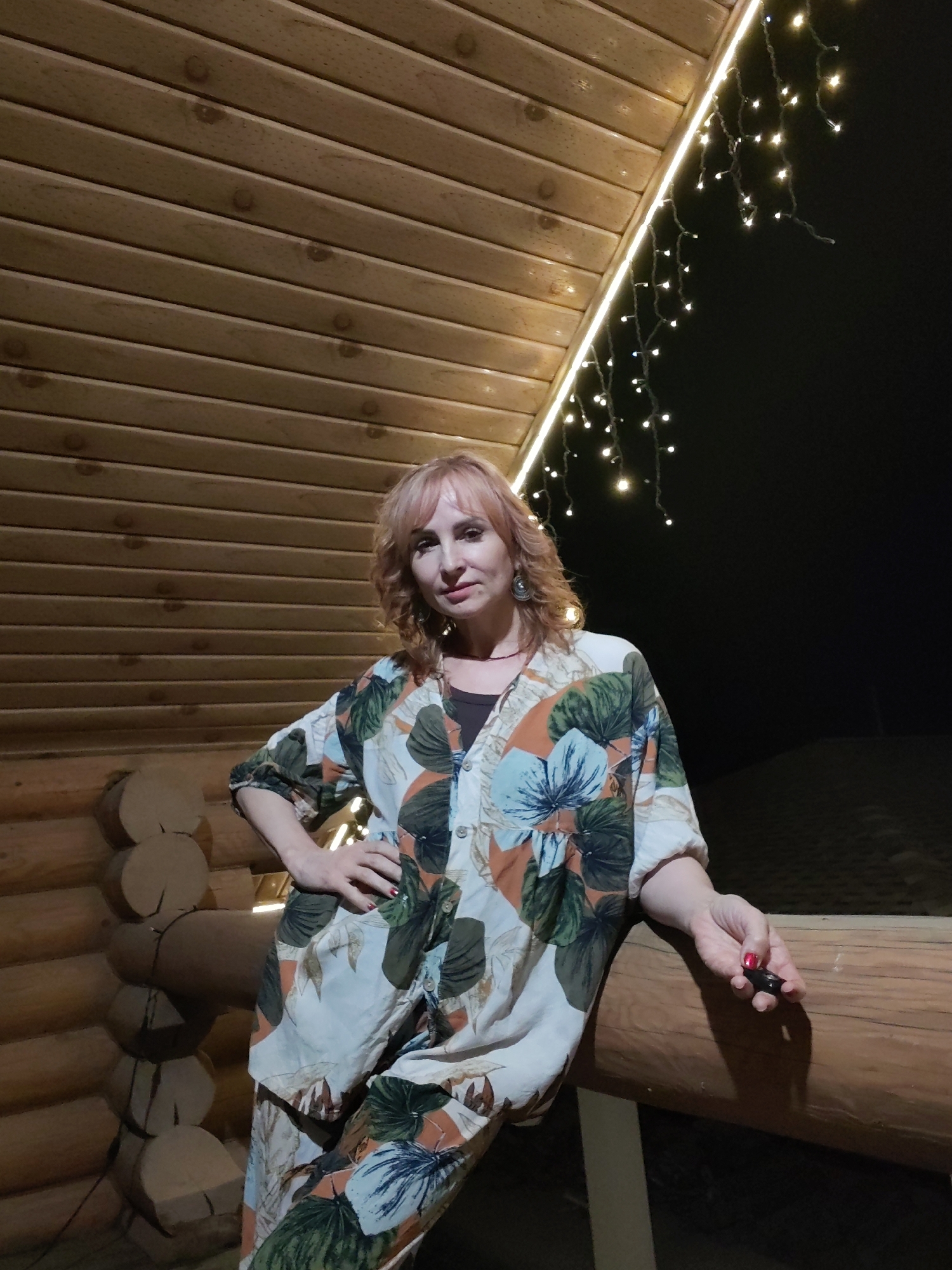 A woman is standing in what appears to be a log cabin, or the exterior of a wooden structure, at night in Старое Михайловское, Russia. The structure has a wooden ceiling and log walls. There are strings of lights hanging along the edge of the ceiling. The woman has light hair and is wearing a floral patterned shirt and pants. She is leaning against a wooden railing with one hand on her hip and the other hand holding something small and dark. The background is dark, suggesting it is night.