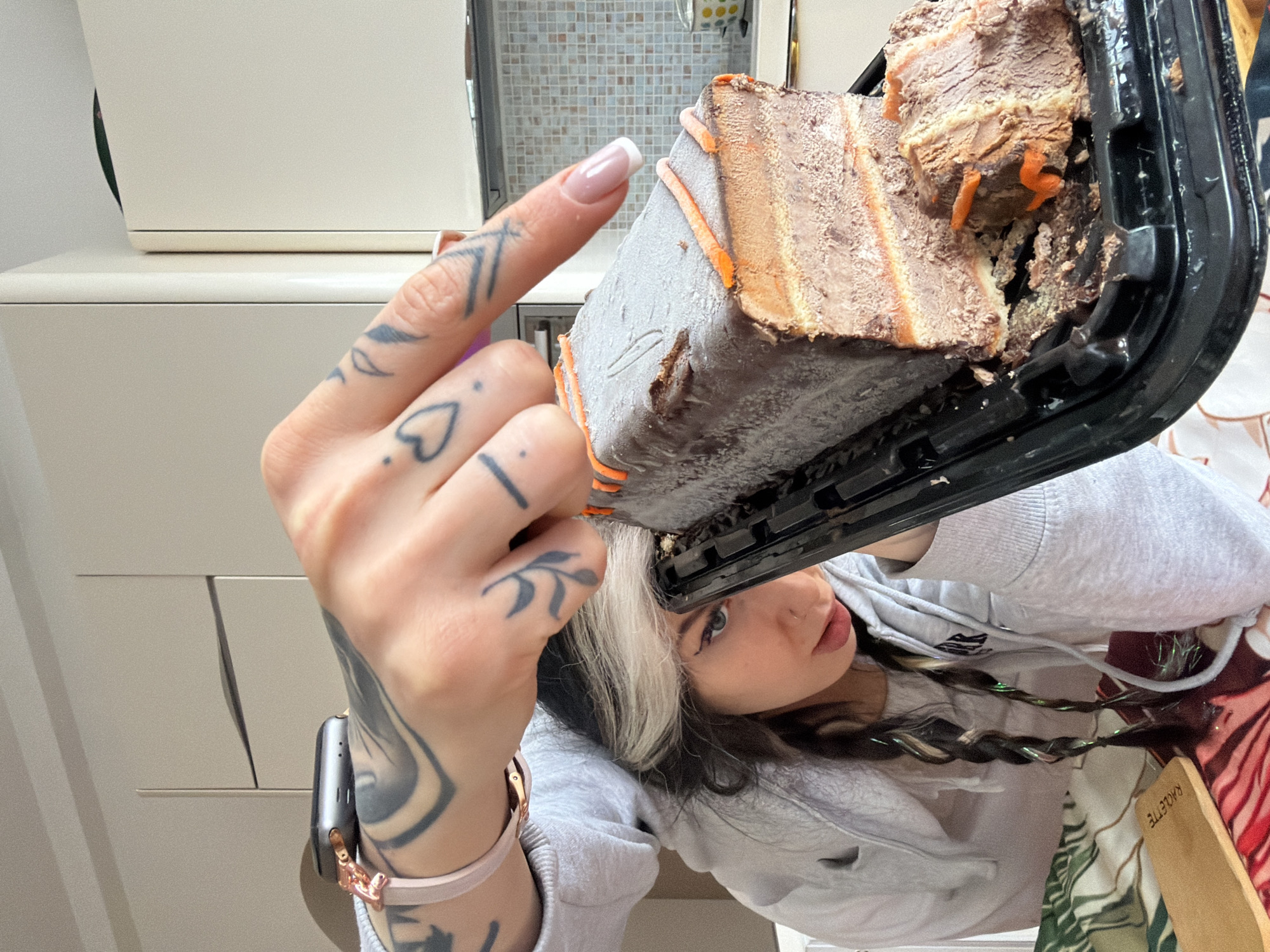 A female subject is depicted in a kitchen setting in Budva, Montenegro, holding a partially consumed frozen chocolate and orange layered dessert. The subject, positioned centrally, has dark hair with a prominent white streak at the front, styled into two braids adorned with green tinsel. She wears a light grey hooded sweatshirt. Her left arm, featuring extensive black ink tattoos, displays an Apple Watch with a rose gold clasp and pink band. Her right hand, also tattooed with a heart symbol on the ring finger and other designs, holds the black plastic tray containing the dessert, with French-tipped nails visible. The dessert, a rectangular loaf, shows layers of brown/chocolate and orange, topped with a light grey frosting and orange decorative lines. A section has been removed from one end, revealing the internal structure and leaving crumbs on the tray. The background consists of light beige kitchen cabinets with vertical metallic handles, and a tiled backsplash made of small, light-colored squares. A dark countertop is visible, along with a partial view of a polka-dotted cup. In the lower-left, a wooden board inscribed with "RACLETTE" rests on a multicolored, patterned tablecloth. The scene is brightly lit.