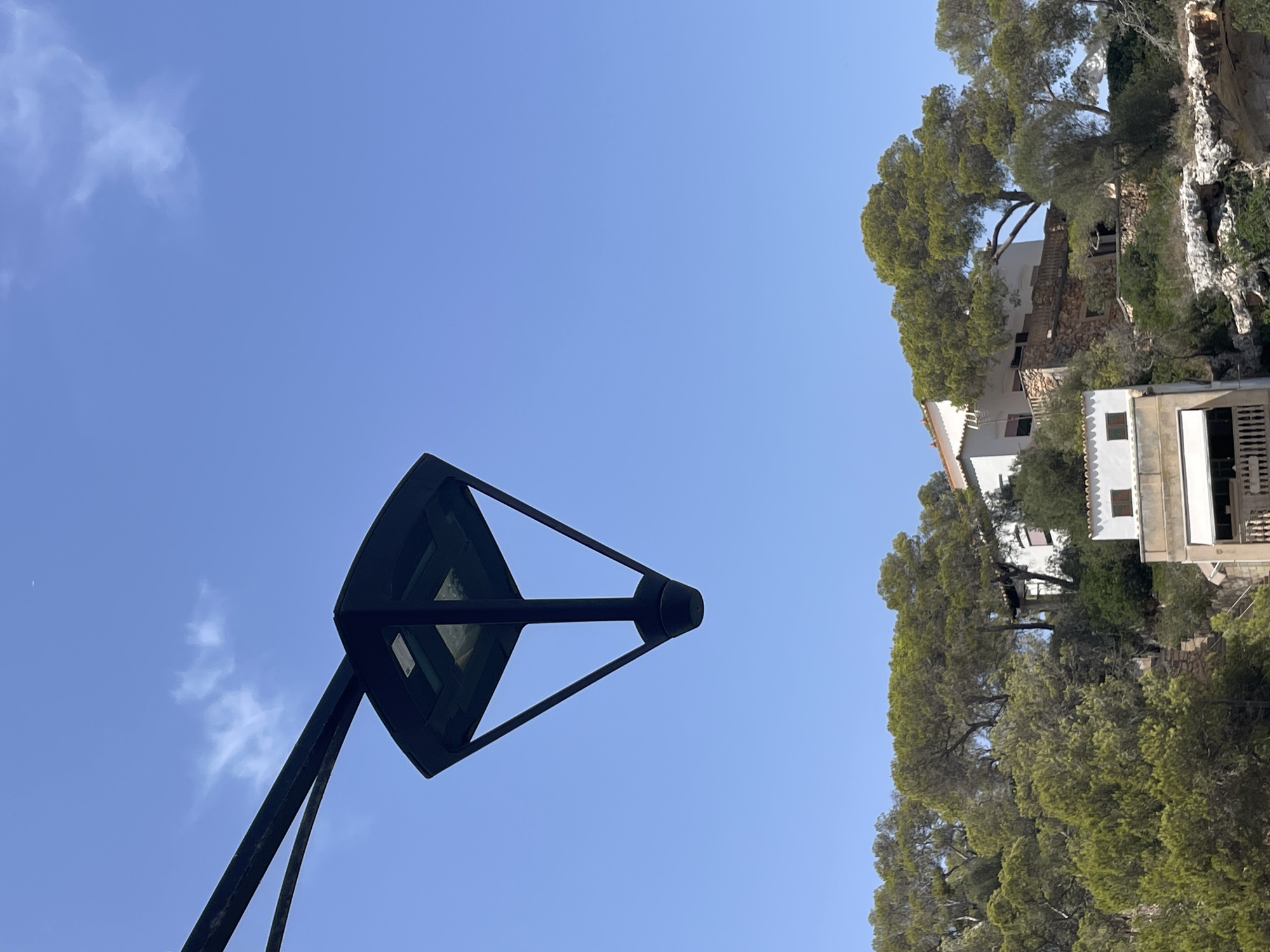 An upward-angle shot shows a dark, modern-style street lamp in the foreground, angled from the left side of the frame, against a bright, clear blue sky. In the background, a hillside is covered with lush green trees and vegetation. Nestled amongst the trees are several white buildings with tiled roofs, suggesting a residential area or possibly a small complex. The architecture of the buildings appears Mediterranean in style, with stone foundations visible on some structures. The overall scene evokes a sunny day in a coastal or hilly region, characteristic of Santanyí, Spain, known for its picturesque landscape and white-washed buildings. There are no people or discernible actions depicted in the image.