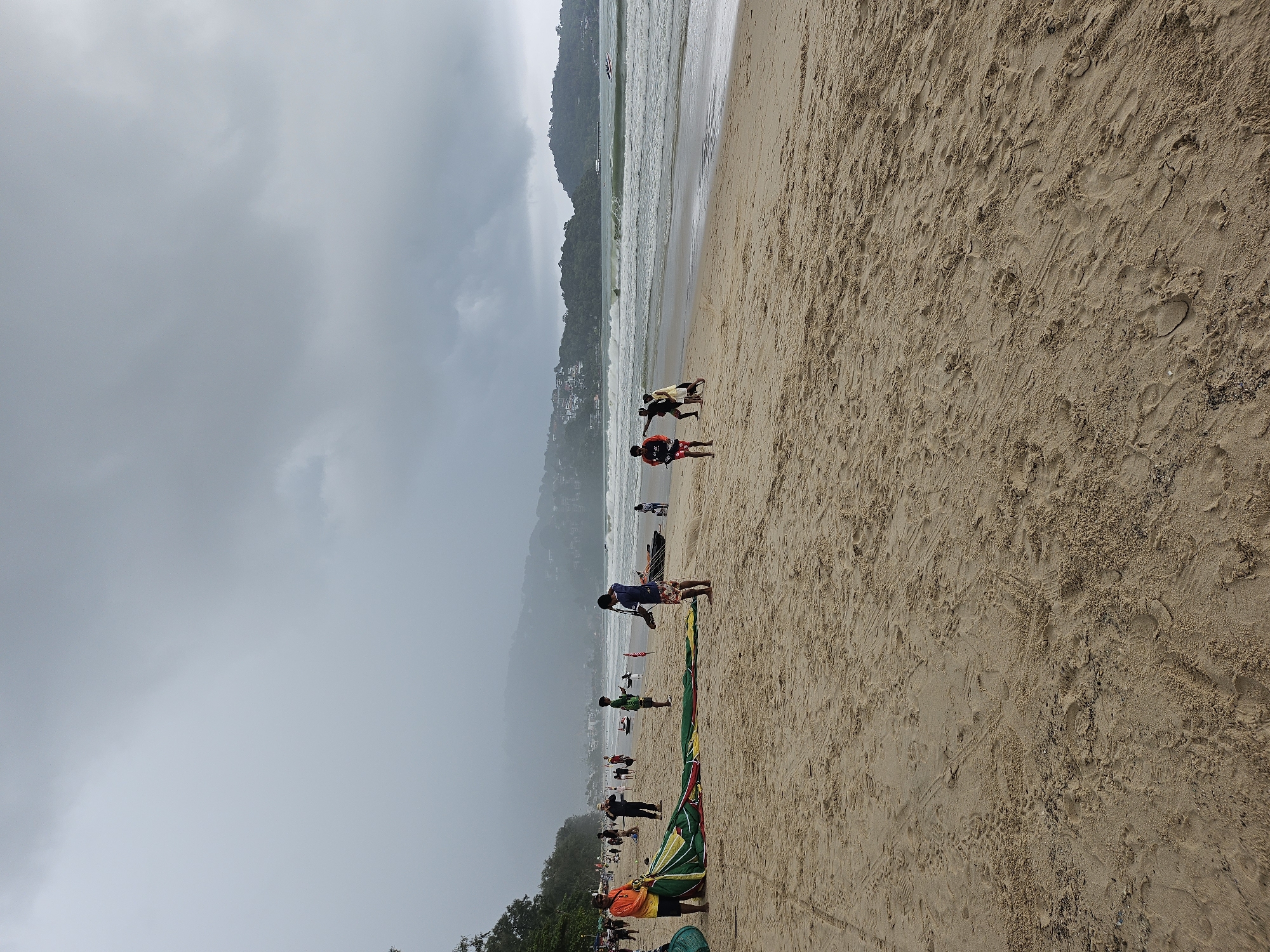 A wide shot of Patong Beach in Phuket, Thailand on an overcast day shows several people on the sandy shore near the ocean. The tide is moderately high with significant waves rolling in. In the foreground, a man in an orange shirt and black shorts is holding a large, folded green and yellow parachute. Nearby, another person in a blue shirt and patterned shorts is also attending to the parachute. Several other individuals are scattered across the beach, some walking and others observing the water. Further in the background, trees and buildings are visible on the hills overlooking the bay. A few small boats are in the water, and a white boat with passengers is visible in the distance. The overall atmosphere is cloudy and humid, suggesting the possibility of rain.