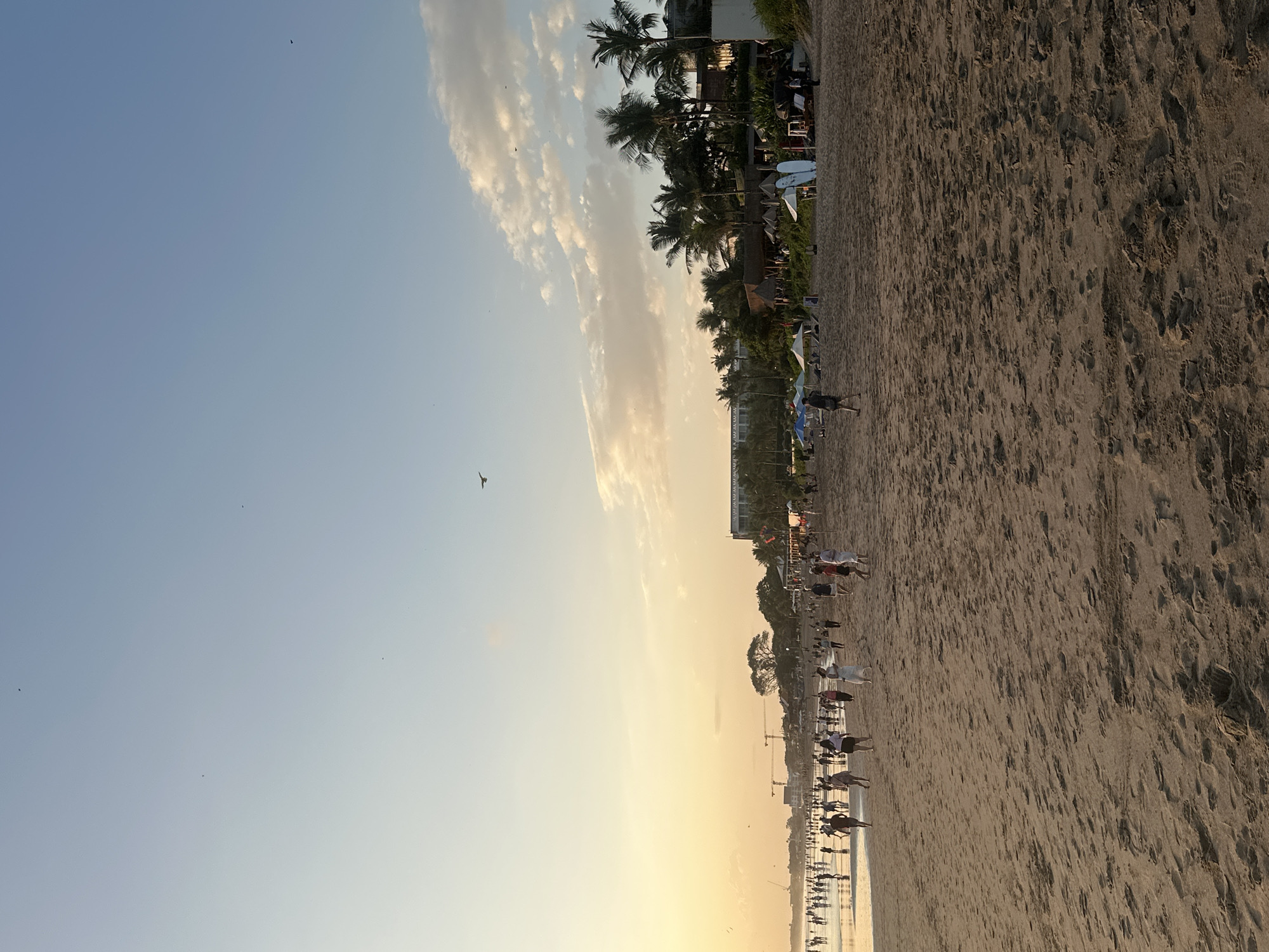 In North Kuta, Indonesia, a beach scene unfolds during what appears to be sunset. The sky is a gradient of pale blue and soft yellow, with scattered clouds and several small birds silhouetted against it. The beach itself is sandy with visible footprints. In the distance, a large crowd of people are walking along the shoreline, some near the water's edge. A line of buildings, including what looks like a hotel with palm trees in front of it, stretches along the coast behind the beachgoers. Beach umbrellas and surfboards are visible in the midground. Two construction cranes are also present in the far distance. The overall atmosphere is one of a public beach at dusk.