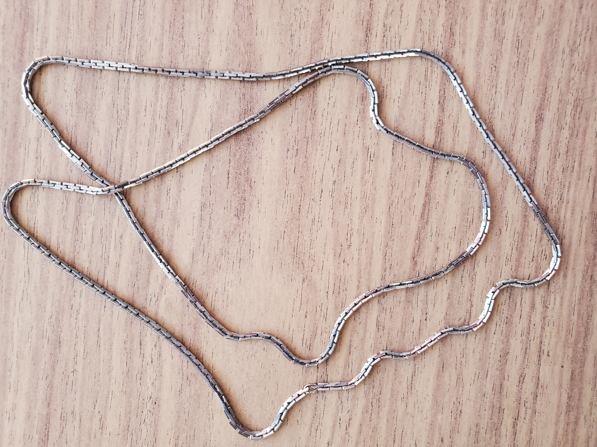 A silver chain necklace lies on a wooden surface, arranged in a loose serpentine pattern. The chain is composed of small, rectangular links connected in a uniform fashion. The wooden surface exhibits a light brown color with visible grain patterns and subtle horizontal lines. The lighting suggests a natural light source, casting soft shadows. There are no people or any other objects present in the image. The specific location in Tiraspol, Moldova, cannot be determined from the provided image.