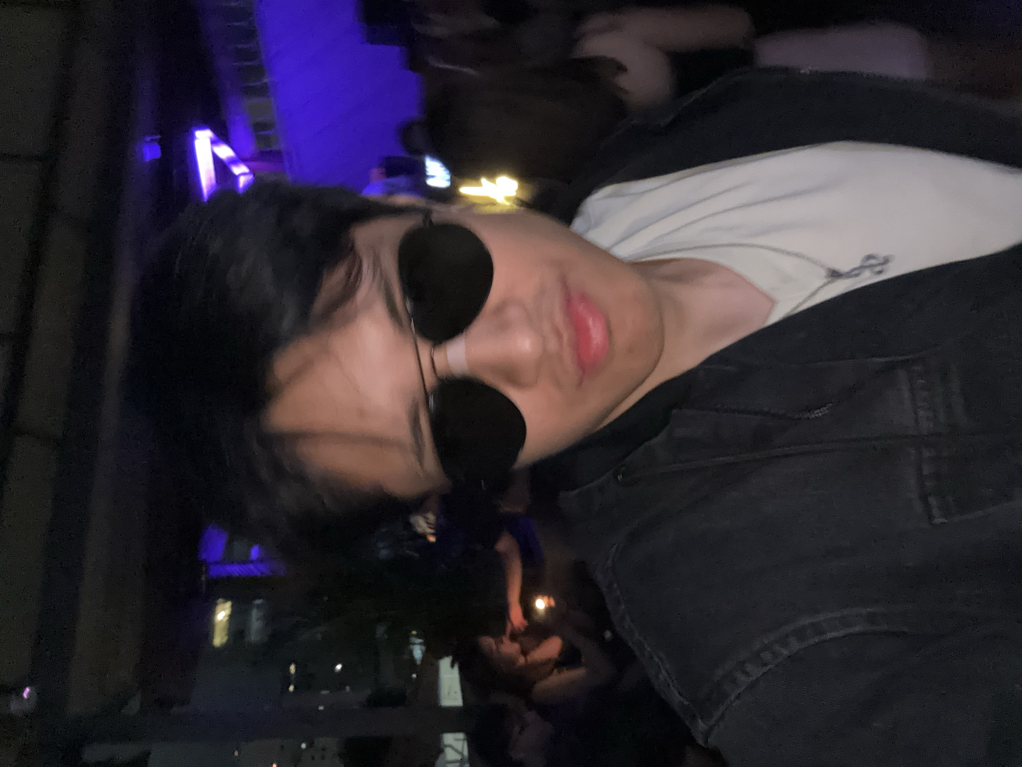 Here is a description of the image:
A selfie taken at night showing a person with dark hair and sunglasses, wearing a white t-shirt underneath a dark jacket. Behind the person, there is a crowd of people and city lights, indicating an outdoor or semi-outdoor location. The location is in São Paulo, Brazil, as suggested by the presence of buildings and urban lights in the background. The purple lighting suggests the setting is at a party.