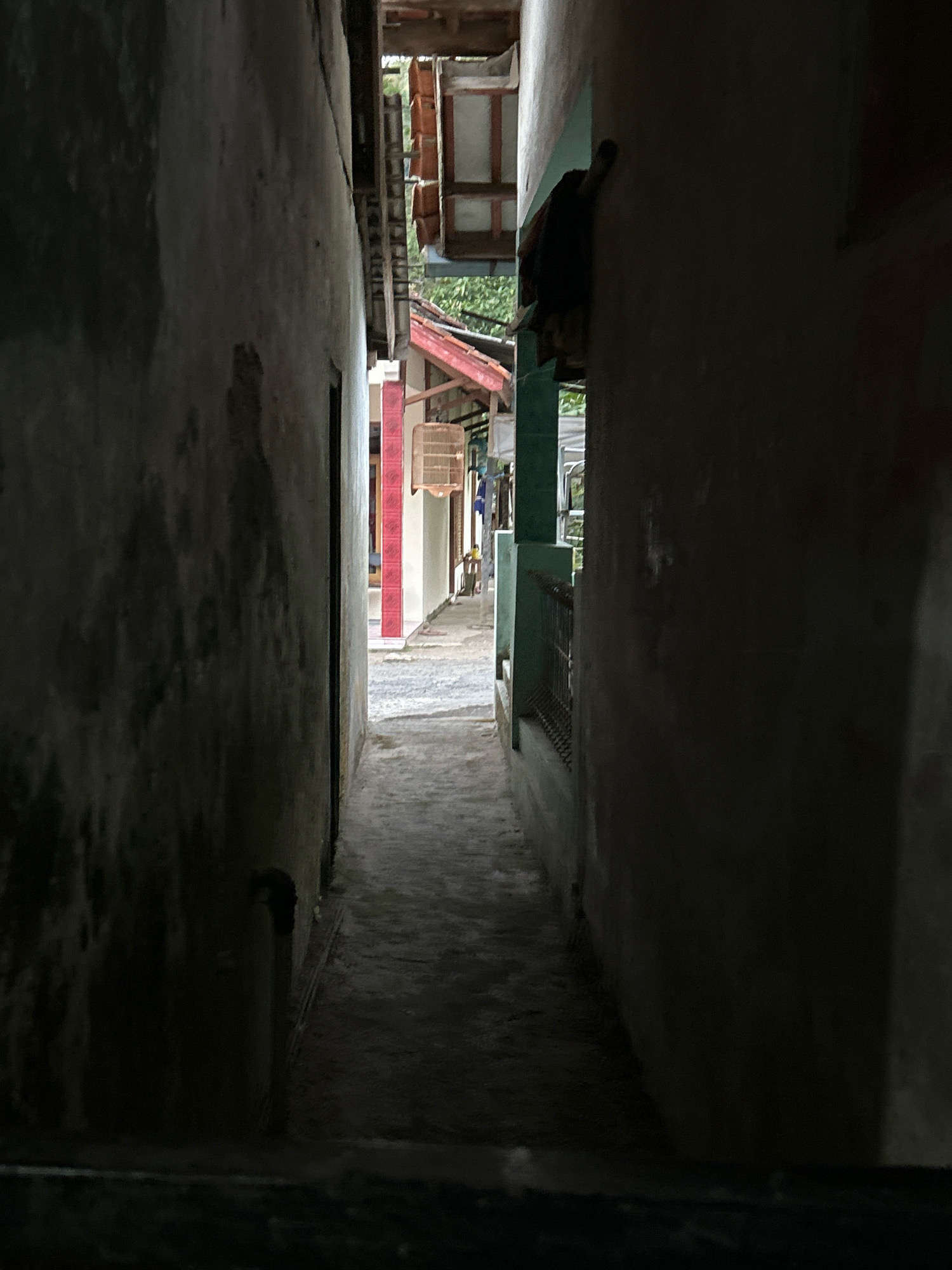The image depicts a narrow alleyway between two weathered buildings. The walls are rough and show signs of age and staining. The alley is paved with concrete and appears to be wet, possibly from recent rain. Sunlight filters in from the opening at the end of the alley, illuminating some of the buildings and structures visible there. These include roofs with red tiles and what appear to be wooden beams. In the distance, there are glimpses of what might be market stalls or shops with brightly colored accents, including red and white striped elements. A birdcage hangs from a structure on the right side of the alley. The scene has a slightly damp and overcast atmosphere. The location is specified as Cihaurbeuti, Indonesia.