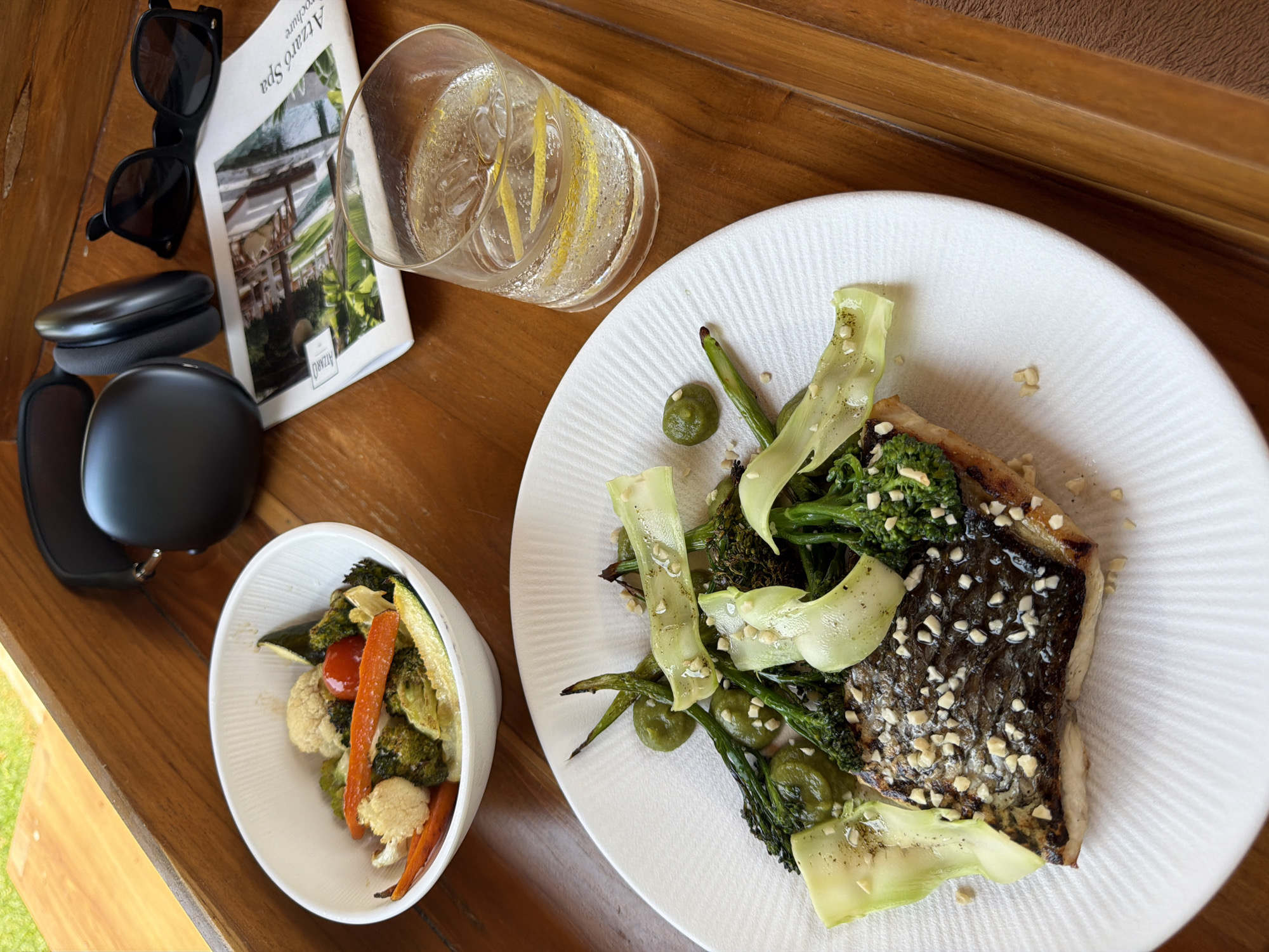 A close-up shot features a white plate with a piece of fish, topped with chopped nuts and served with green vegetables, including broccolini, asparagus, and bok choy ribbons. To the left, a white bowl contains a medley of roasted vegetables such as carrots, cauliflower, broccoli, and zucchini. In the background, a glass of sparkling water with lemon slices sits next to a brochure for "Azaro Spa." A pair of black headphones and black sunglasses rest on a wooden surface, suggesting a relaxed setting. The overall scene is set on a wooden tray. The lighting indicates it's daytime, possibly outdoors given the hint of greenery visible in the upper left corner. The composition implies a light meal or snack, possibly enjoyed during a break or while relaxing.