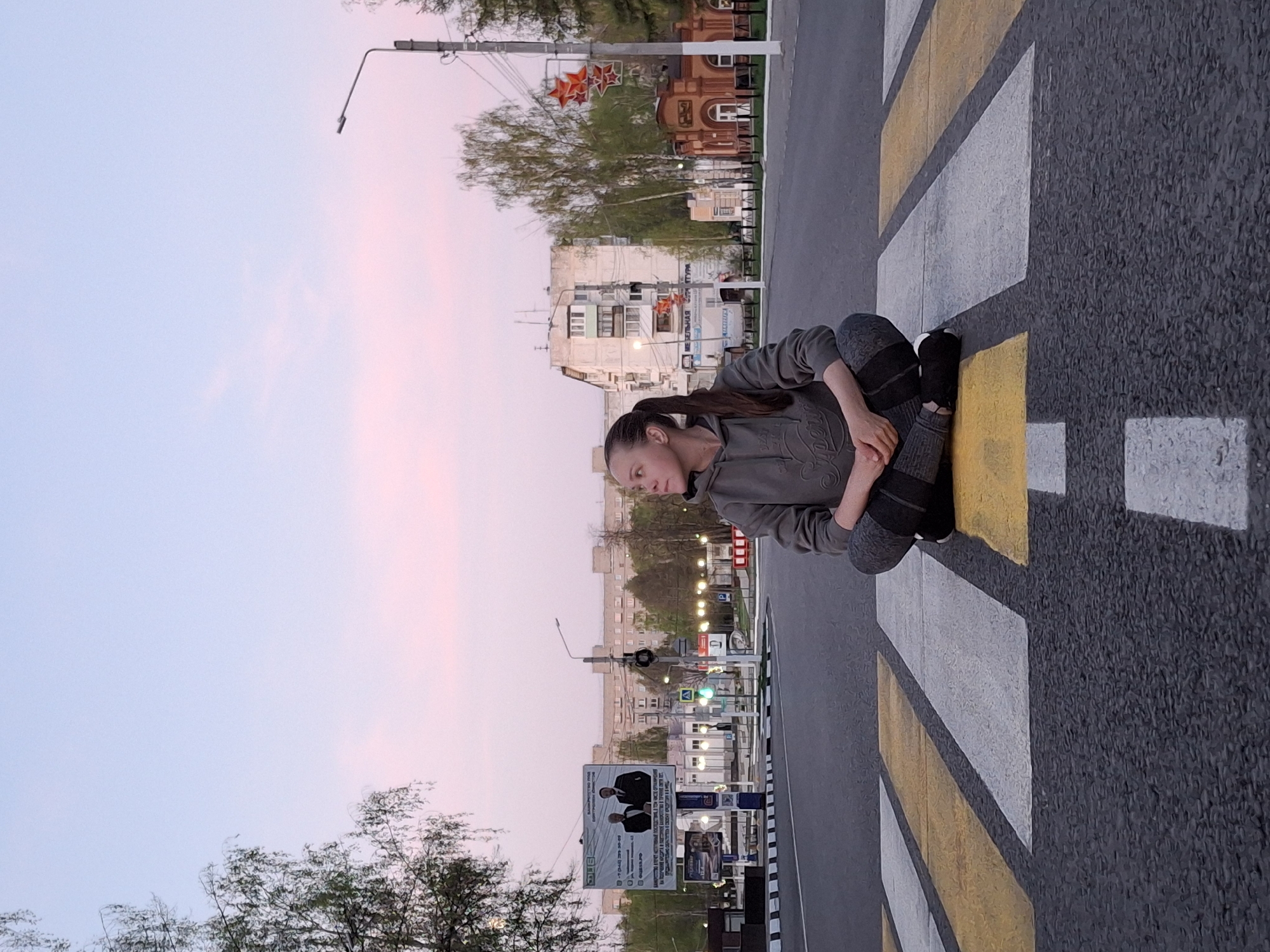 A woman is sitting cross-legged on a crosswalk in the city of Chaikovsky, Russia. The crosswalk has alternating white and yellow stripes. The woman has long brown hair pulled back into a ponytail. She is wearing a gray sweatshirt and gray leggings with black socks and shoes. The background shows a city street with buildings, trees, streetlights, and billboards. The sky is light blue with a hint of pink.