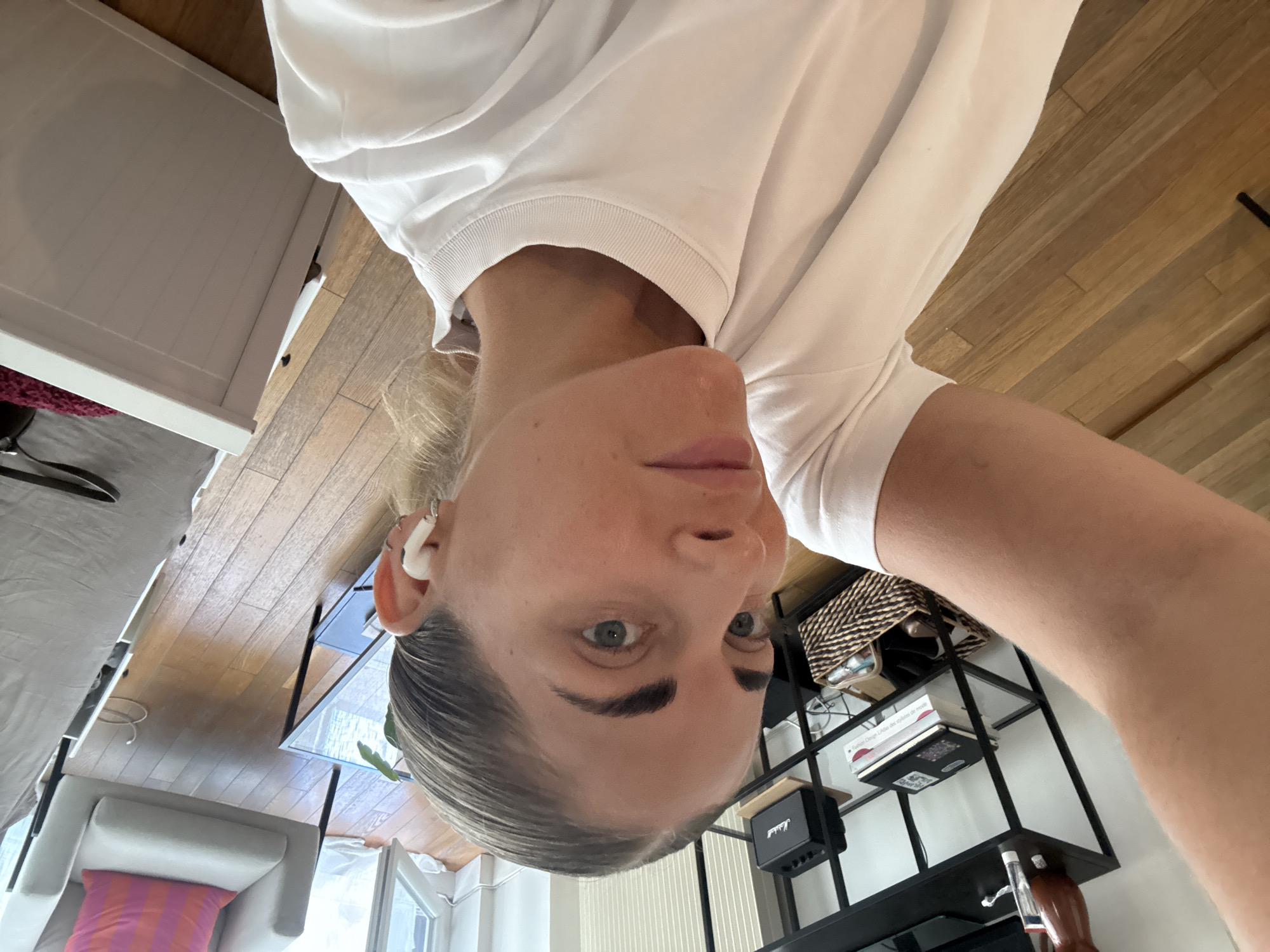 The image shows a close-up, selfie-style shot of a woman in Paris, France. She is wearing a white t-shirt and has blonde hair pulled back. She is also wearing white wireless earbuds. The background includes a wooden floor, a black shelf unit with books and a speaker, a glass-topped table, and a light-colored armchair with a pink and orange striped pillow. A partially visible white dresser is also in the background. The scene appears to be in a residential setting.