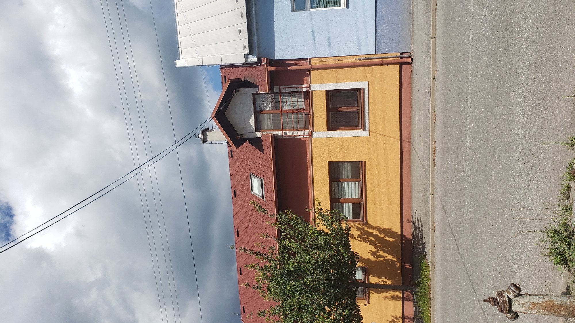 The image shows a street scene in Prešov, Slovakia, featuring a two-story building with an orange facade and a red roof, situated next to a light blue building with a white roof. The orange building has several windows, a small balcony on the upper floor, and a tree with dappled shadows on its facade. Power lines are visible stretching across the sky. In the foreground, a rusty fire hydrant stands beside an asphalt road and a patch of grass. The sky is partly cloudy with dark storm clouds gathering.