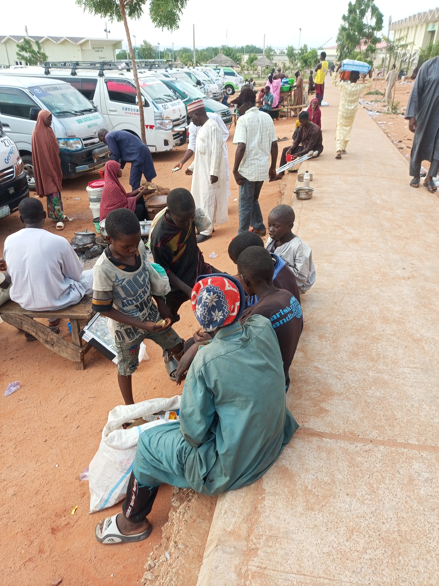 The image depicts an outdoor scene in Gombe, Nigeria, likely near a transportation hub or market. Several white minibuses are parked in the background, some with branding that appears to be related to local government or transportation services. A group of people, predominantly men and boys, are gathered in the foreground and midground. Some are seated on the ground or on a wooden bench, while others are standing or walking.
The individuals are dressed in traditional African attire, including flowing robes and head coverings. One man in a white robe is interacting with a woman in a reddish-brown hijab who is kneeling near some cooking pots and food items, suggesting a communal meal or food preparation. Several young boys are clustered together, some appearing to be eating or engaged in conversation. One boy in a grey shirt is holding a piece of food. Another boy is wearing a dark shirt with blue lettering. A man in a light-colored robe is walking away from the group towards the background.
The ground is covered in reddish-brown soil, with a concrete walkway running through the scene. Scattered items include plastic bags, a white sack, cooking pots, and containers. The environment suggests a somewhat dusty and open area, possibly with some trees and low buildings in the distance. The overall impression is one of daily life and community interaction in an urban setting.