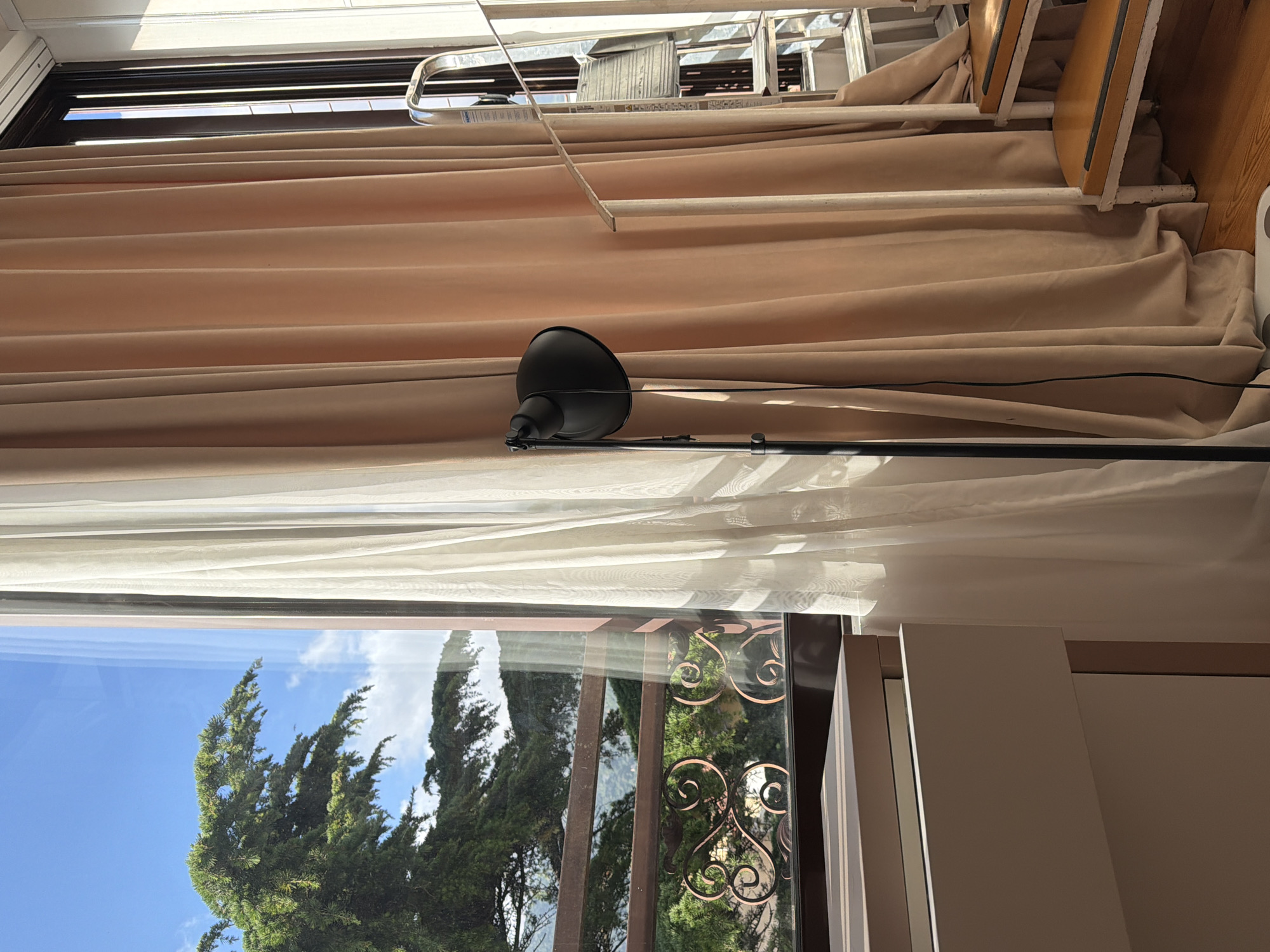 The image depicts an interior view through a window. Outside, there are green trees visible under a blue sky with some clouds. Inside, there is a dark-colored lamp standing in front of the window. The window has a decorative metal design. Beige-colored curtains are behind the lamp. Above the curtains, there is a silver-colored ladder resting on what appears to be a wooden structure. The scene appears to be inside a room in a building. There is not enough information to confirm the location within the city of Budva, Montenegro.