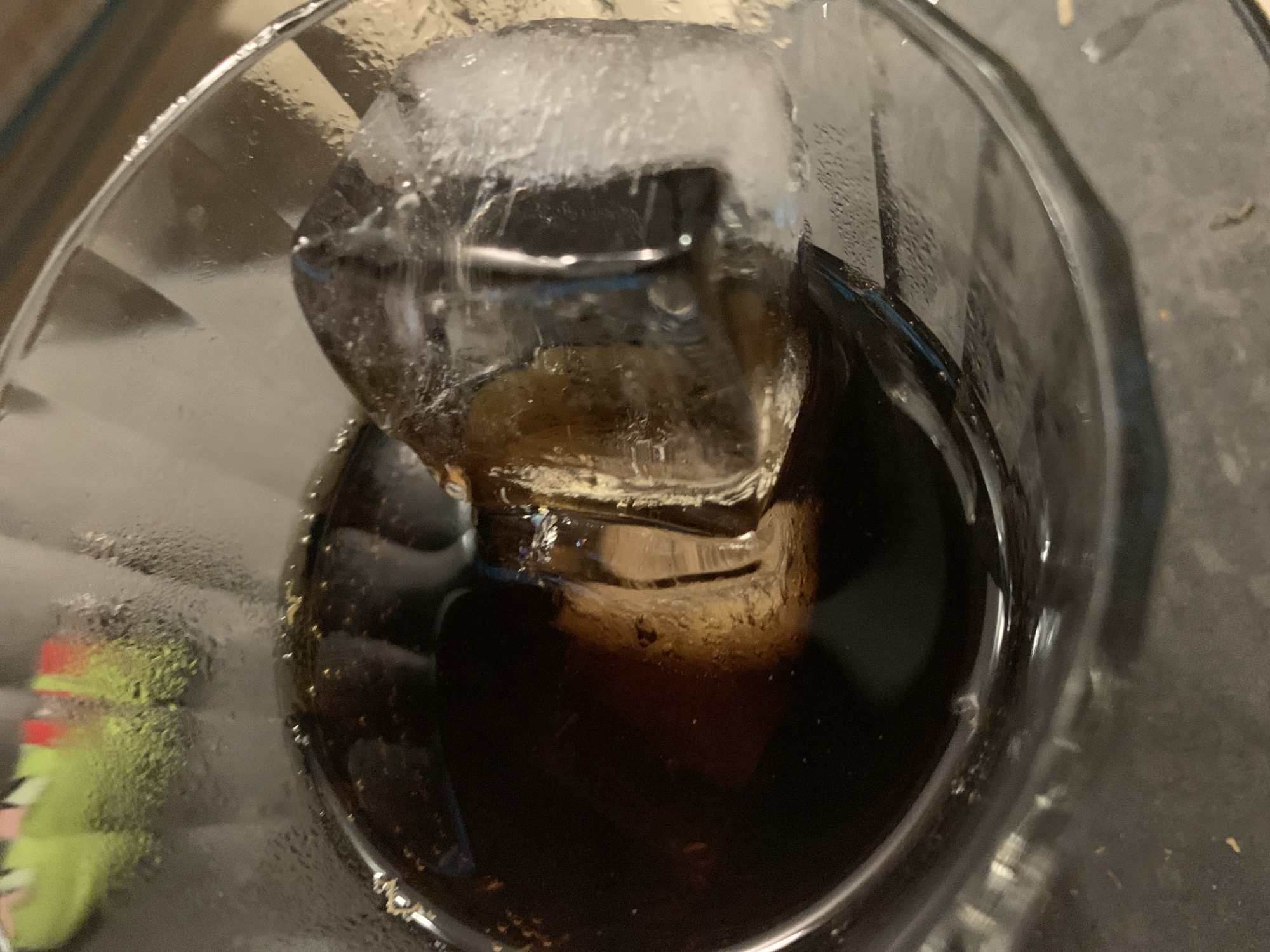 The image provides a close-up, high-angle view looking directly into a glass containing a dark beverage and ice cubes. The clear glass exhibits significant condensation and small water droplets on its exterior, indicating a cold drink.
Two large, irregularly shaped ice cubes are prominent, floating in the dark liquid. The ice shows a slightly frosty and opaque texture on its upper surface, becoming clearer where it is submerged. Reflections of light are visible on the ice.
The beverage itself is a deep, opaque dark brown or black, consistent with cola or a similar carbonated soft drink. Small bubbles are visible on the liquid's surface and along the edges of the ice cubes, suggesting carbonation.
In the blurred background to the left of the glass, an out-of-focus, brightly colored object is partially visible. It appears to be a green item with red and white markings, possibly a toy or a cartoon character. The glass rests on a dark, slightly textured surface.
No people are visible in the image. There is no discernible text present. The scene depicts a simple, everyday moment of a cold drink, likely indoors. While the context of Tel-Aviv, Israel is provided, there are no specific visual cues within the image to confirm this location.