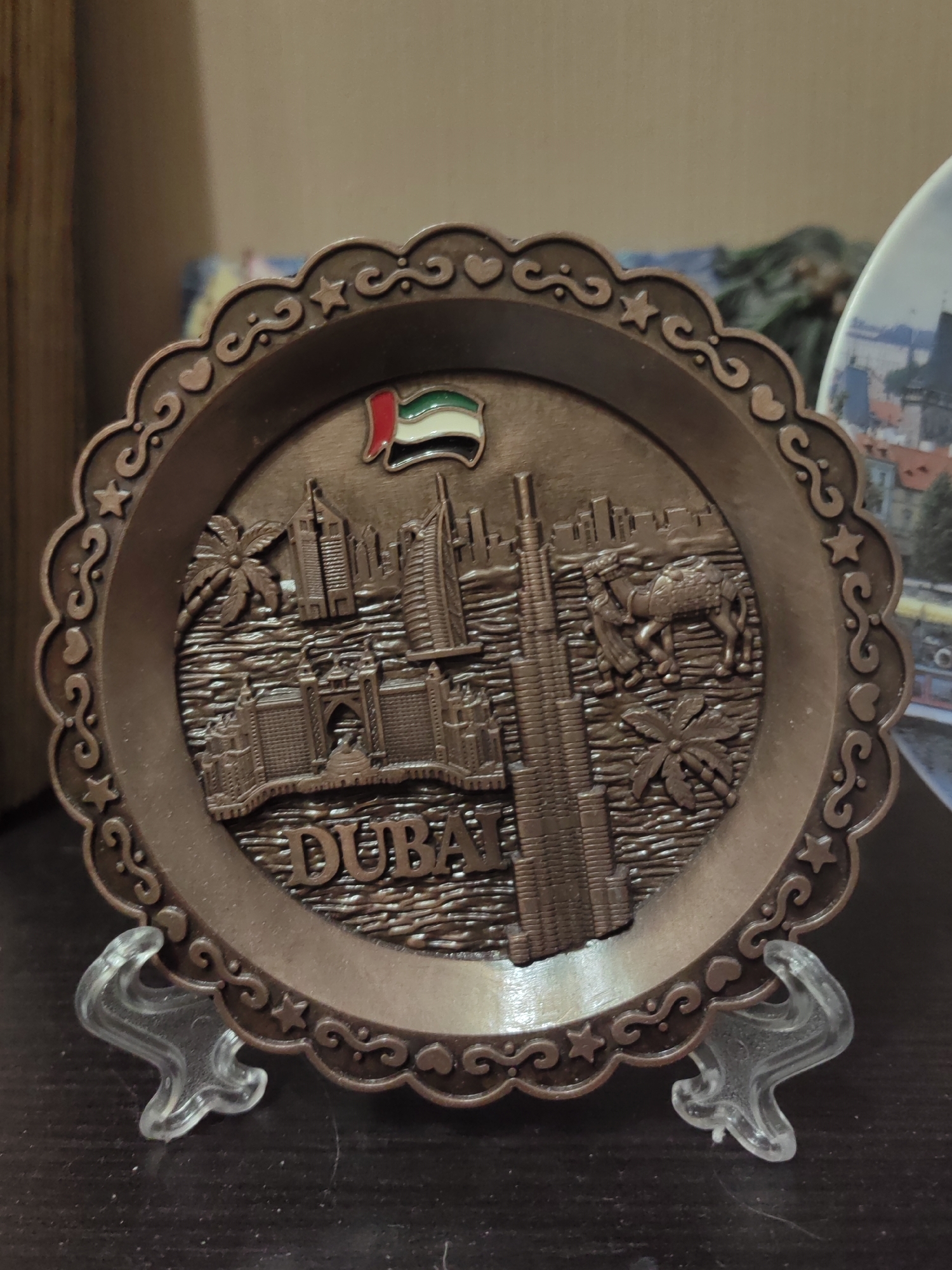 The image shows a close-up, eye-level shot of a bronze-colored decorative plate featuring a relief of Dubai's skyline. The plate has a scalloped edge with a repeating pattern of hearts and swirls, and a star within some of the swirls. At the top center of the plate is a small enamel pin of the United Arab Emirates flag. The relief depicts several skyscrapers, including what appears to be the Burj Khalifa, a palm tree, a camel with a rider, and a building resembling the Burj Al Arab. The word "DUBAI" is embossed in large, blocky letters across the bottom of the relief. The plate is supported by two clear plastic stands. The background is blurred but suggests an indoor setting with wooden furniture and other decorative items. The image does not show the city of Ostafievo, Russia.