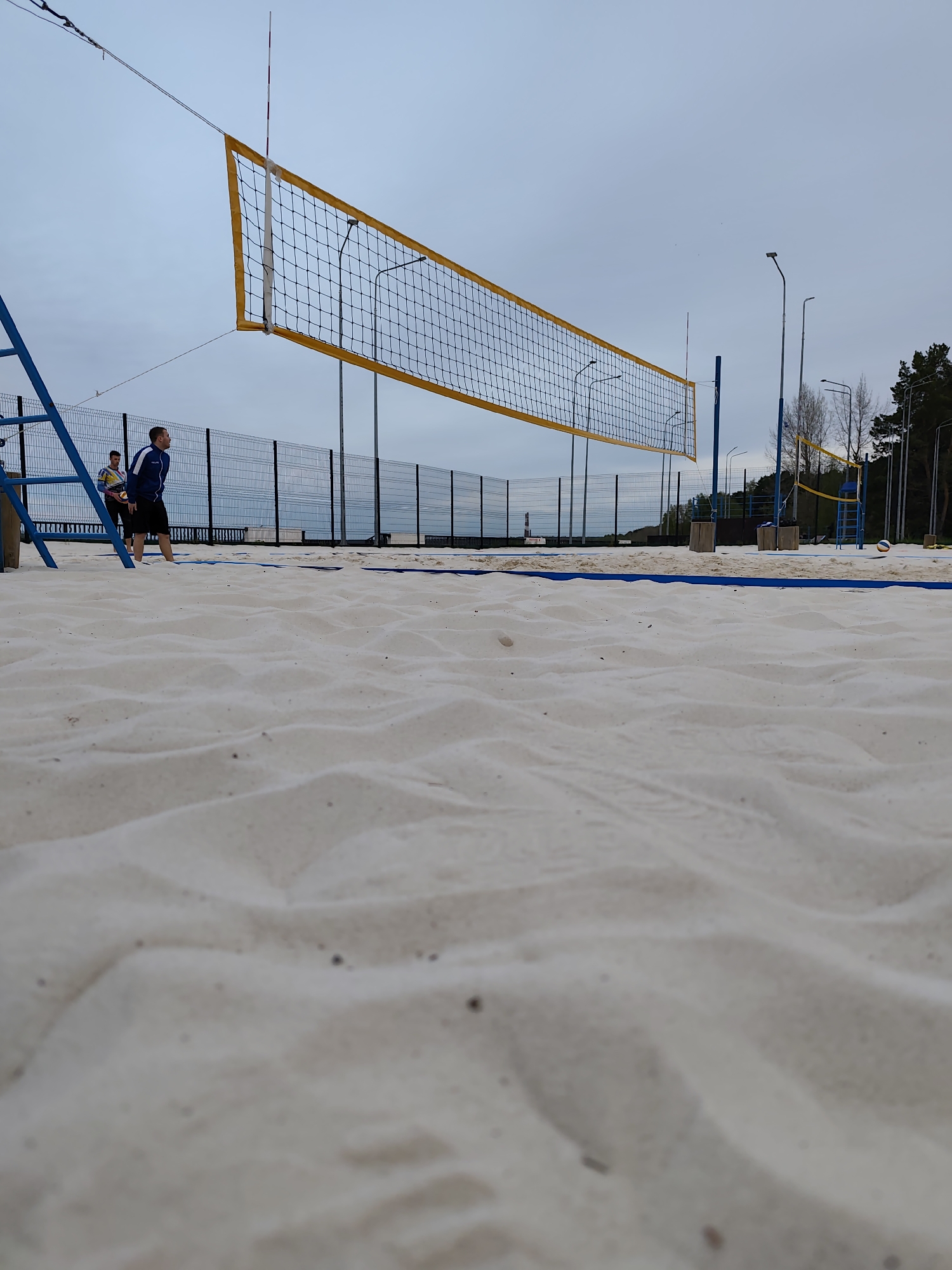 A volleyball court is depicted in a ground-level shot. The court is filled with white sand, its surface marked by footprints and patterns. A blue border marks the boundaries of the court. A net with a yellow top runs across the middle. Two men stand near a blue structure on the left side of the image, suggesting they may be playing volleyball. Streetlights and trees line the background, as well as a tall fence. In the distance, another volleyball net and ball are visible. The sky is overcast and grey. This is likely the Chaikovsky beach volleyball court, Russia.