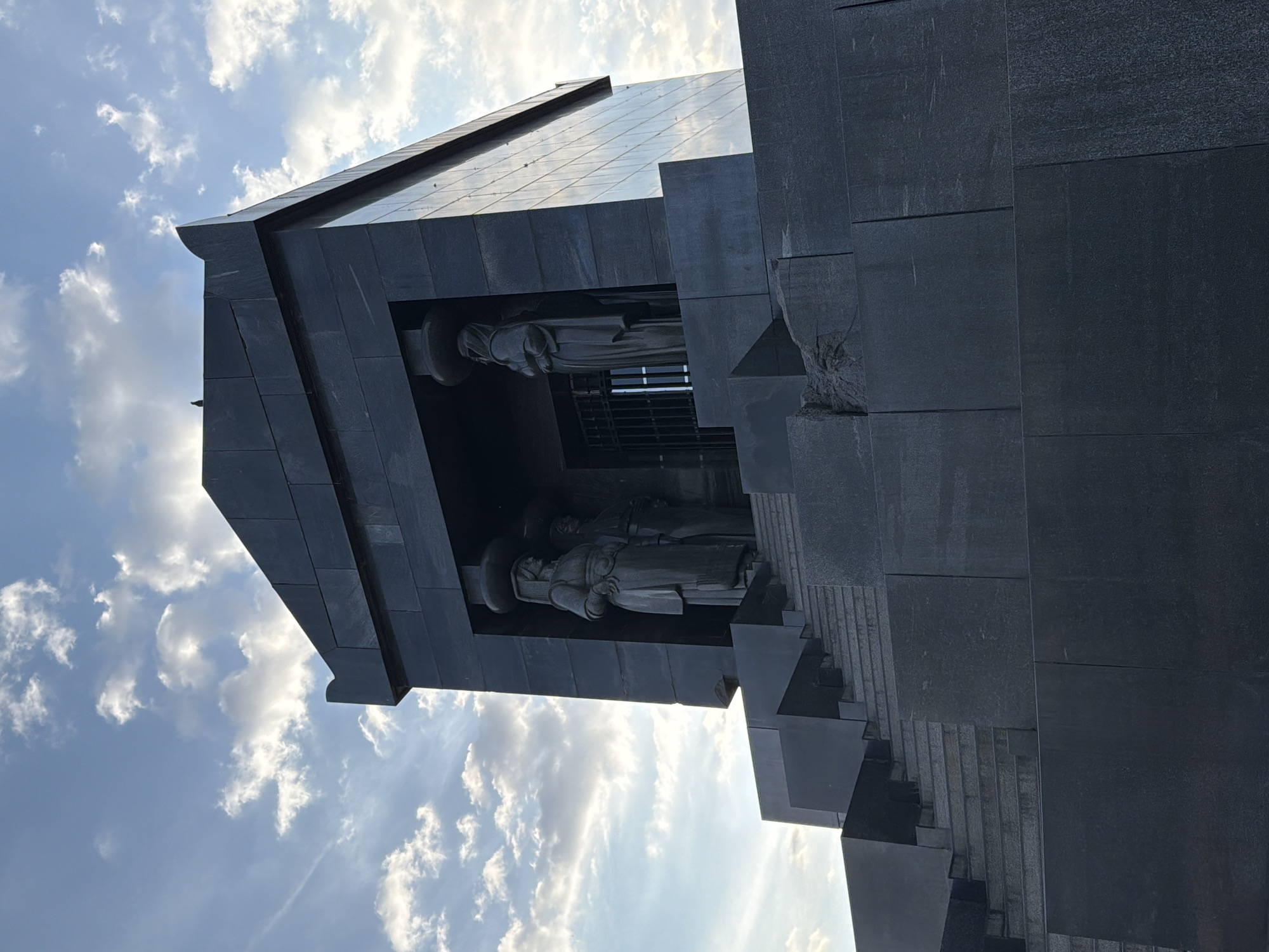 The image shows the Monument to the Defenders of Belgrade in Vozdovac Urban Municipality, Serbia. The monument is constructed of dark stone and features two figures placed inside of the structure. The sky is visible in the background, and some clouds are apparent.