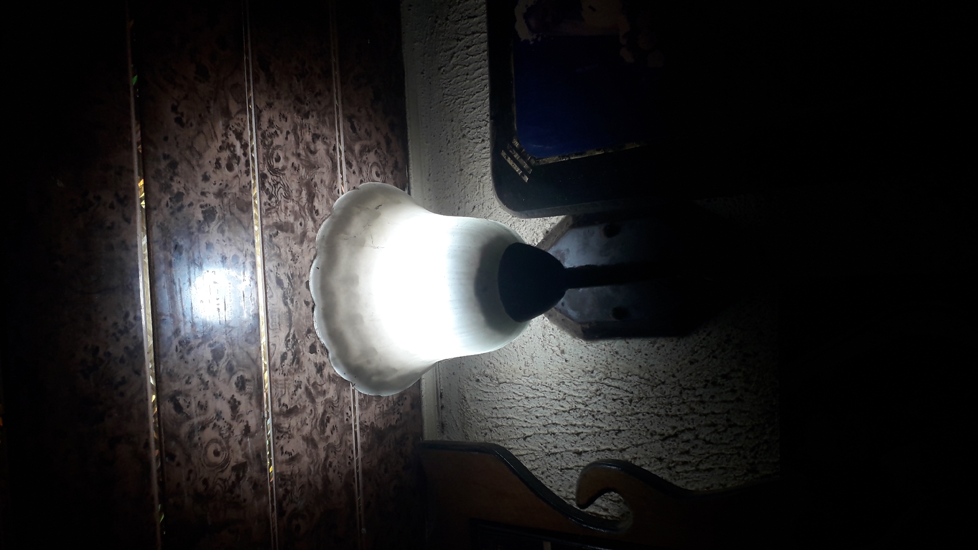 The image shows an illuminated light fixture attached to a wall. The light emits a bright glow that reflects on a nearby textured wall. Adjacent to the light is a dark, mottled surface, possibly a wall panel or decoration. Below the light, part of a wooden structure or furniture piece is visible. Due to the darkness of the image, other details of the room and any potential activity occurring in the scene are obscured. The city and its location of Nkpor, Nigeria were provided by the user and not observable from the visual data.