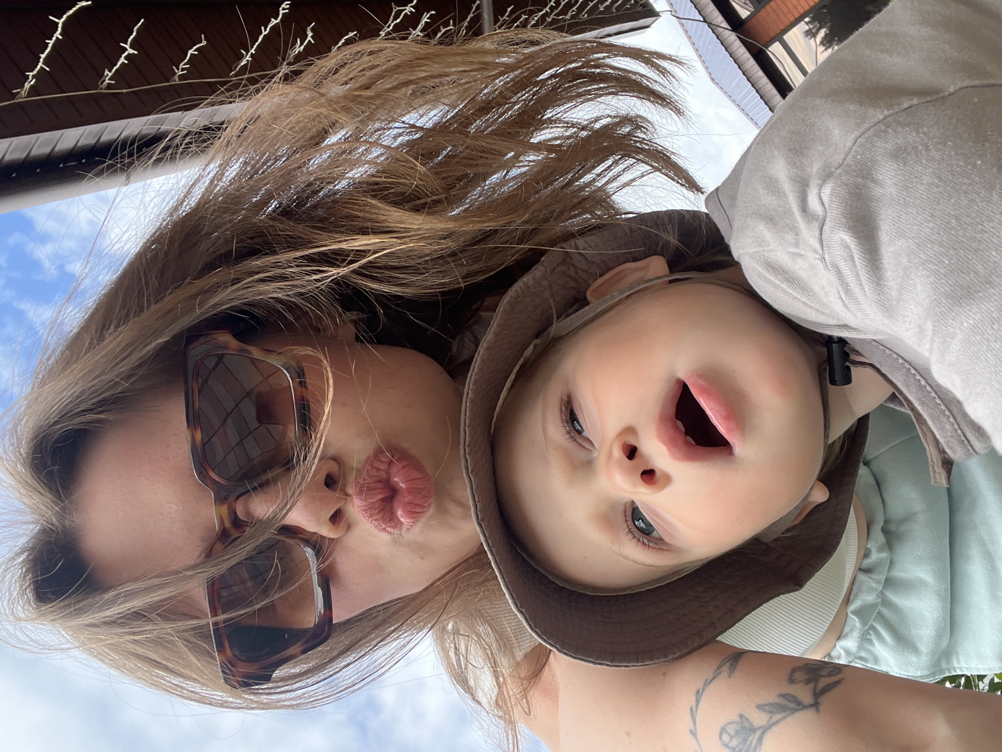 An upward-angled outdoor selfie features an adult and a young child. The adult, positioned in the upper frame, has light brown hair, wears tortoiseshell-framed sunglasses, and pouts her lips for a kiss. A black tattoo depicting floral or leafy branches is visible on her left upper arm. She wears a light-colored ribbed top and what appears to be light green shorts or pants. Below her, a young child, likely a toddler, wears a brown bucket hat and a light grey long-sleeved shirt. The child has light blue eyes, light hair beneath the hat, and an open mouth, looking upwards and to the right. The background consists of a blue sky with scattered white clouds, and elements of a building are visible in the upper right. These elements include a dark brown overhang or soffit and a light-colored, possibly corrugated metal roof, beneath which a string of white decorative lights is strung. There is no visual information within the image to confirm the location as the city of Chaikovsky, Russia.