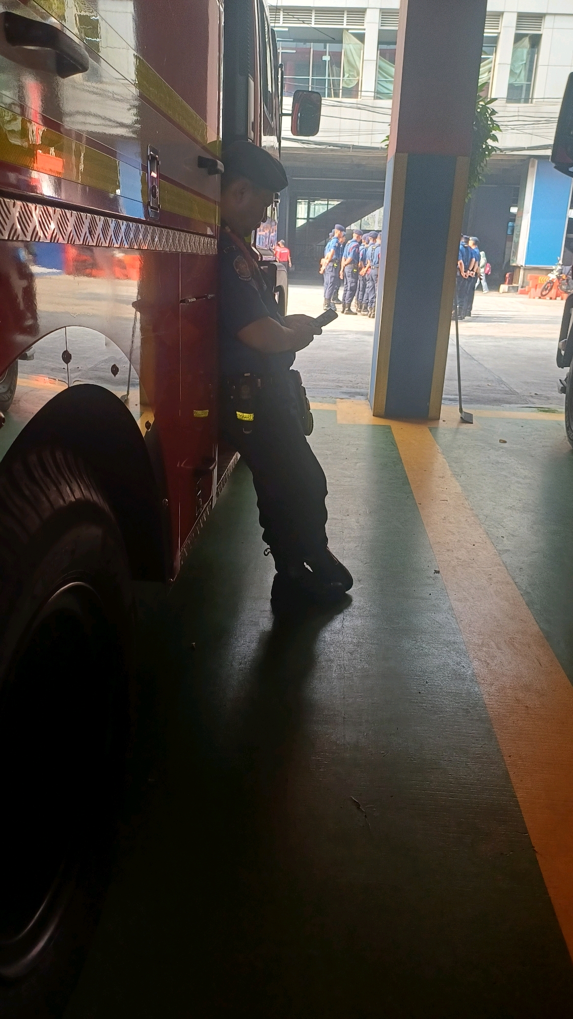 A man in a dark blue uniform, complete with a beret and tactical belt, leans against the side of a red fire truck. He is looking down at a mobile phone in his hands, with his body angled away from the camera. The scene appears to be an outdoor parking area or bay, with a concrete floor marked with yellow lines. In the background, several other individuals in similar uniforms are standing in formation. The architecture in the background suggests a modern building complex, possibly a public facility or administrative center. The lighting indicates it is daytime. The overall impression is one of a momentary pause or downtime for the personnel and the emergency vehicle.