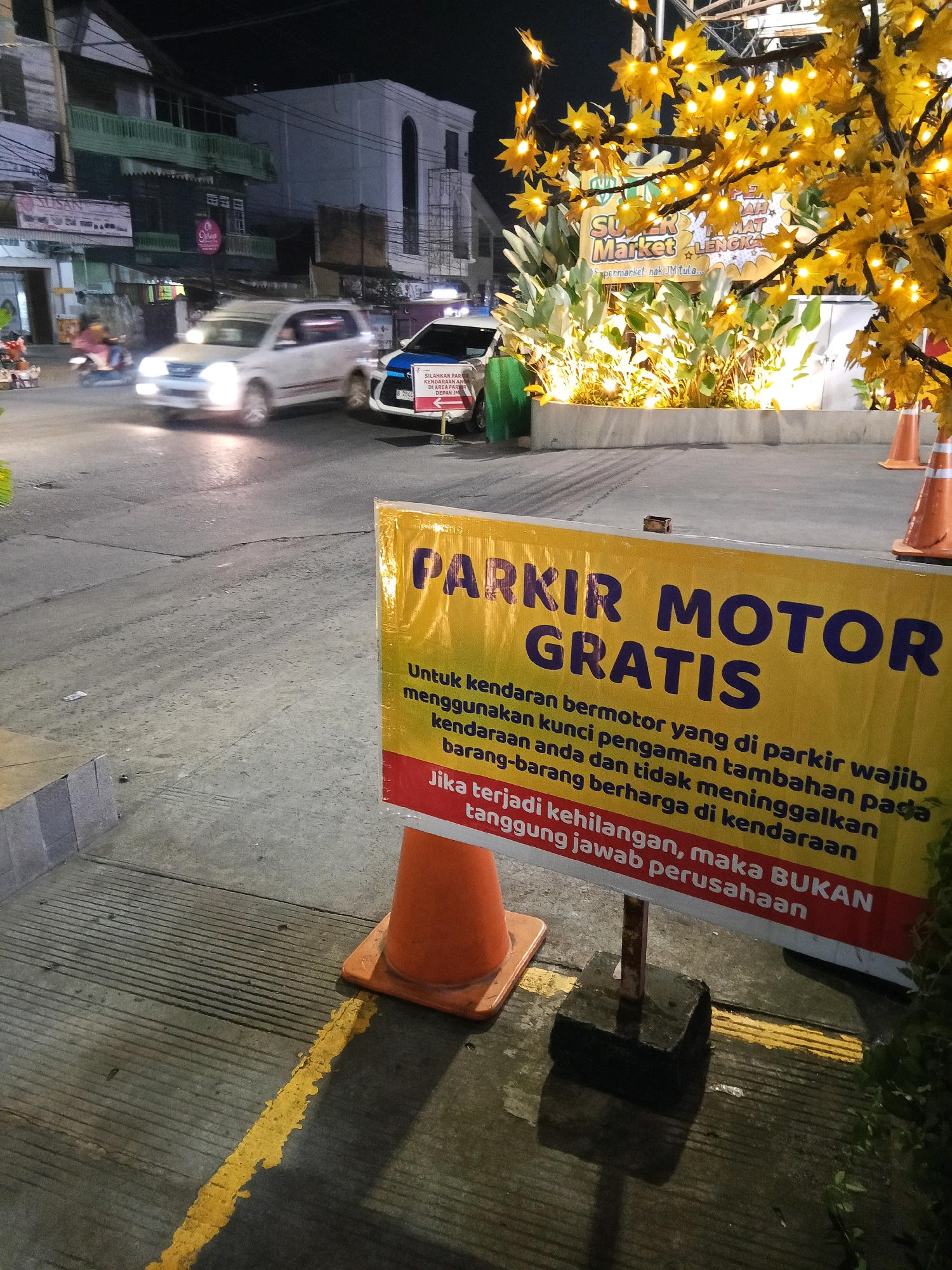 The image depicts an outdoor scene at night, likely a street or parking area in Lubuklinggau, Indonesia. A prominent yellow and red sign in the foreground reads "PARKIR MOTOR GRATIS" (Free Motorcycle Parking) and provides instructions regarding vehicle security. Behind the sign, a paved surface with yellow markings is visible, along with orange traffic cones. In the background, a white vehicle, possibly an SUV, is moving, with its headlights illuminated. Another vehicle, identified as a police car by its markings, is stationary. The scene is illuminated by artificial lights, including string lights on a decorative tree with yellow foliage, and lights from buildings in the background. The environment appears to be commercial or urban, with buildings lining the street.