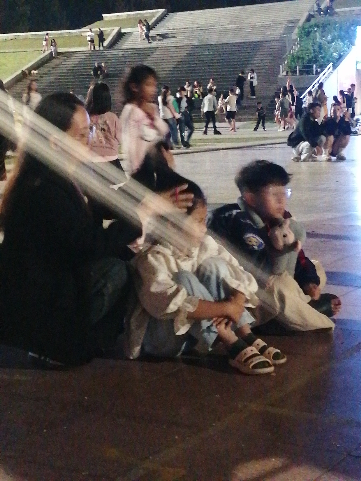 The image captures a group of people in an outdoor setting with tiered seating in the background. In the foreground, several individuals are seated on the ground. To the left, a person in dark clothing is partially visible. Beside them, a child in a light-colored outfit with blue pants is seated. To the right of the child, a boy wearing a dark jacket over a lighter shirt is holding a stuffed animal. The environment appears to be a public space, possibly an amphitheater or plaza, with concrete ground and artificial lighting. The background shows a large, stepped structure with people gathered on different levels, some appearing to be walking or standing. The scene is consistent with a public gathering or event. This location is likely the Da Lat Flower Park, specifically near the amphitheater area, given the architecture and the presence of numerous people in a recreational setting.