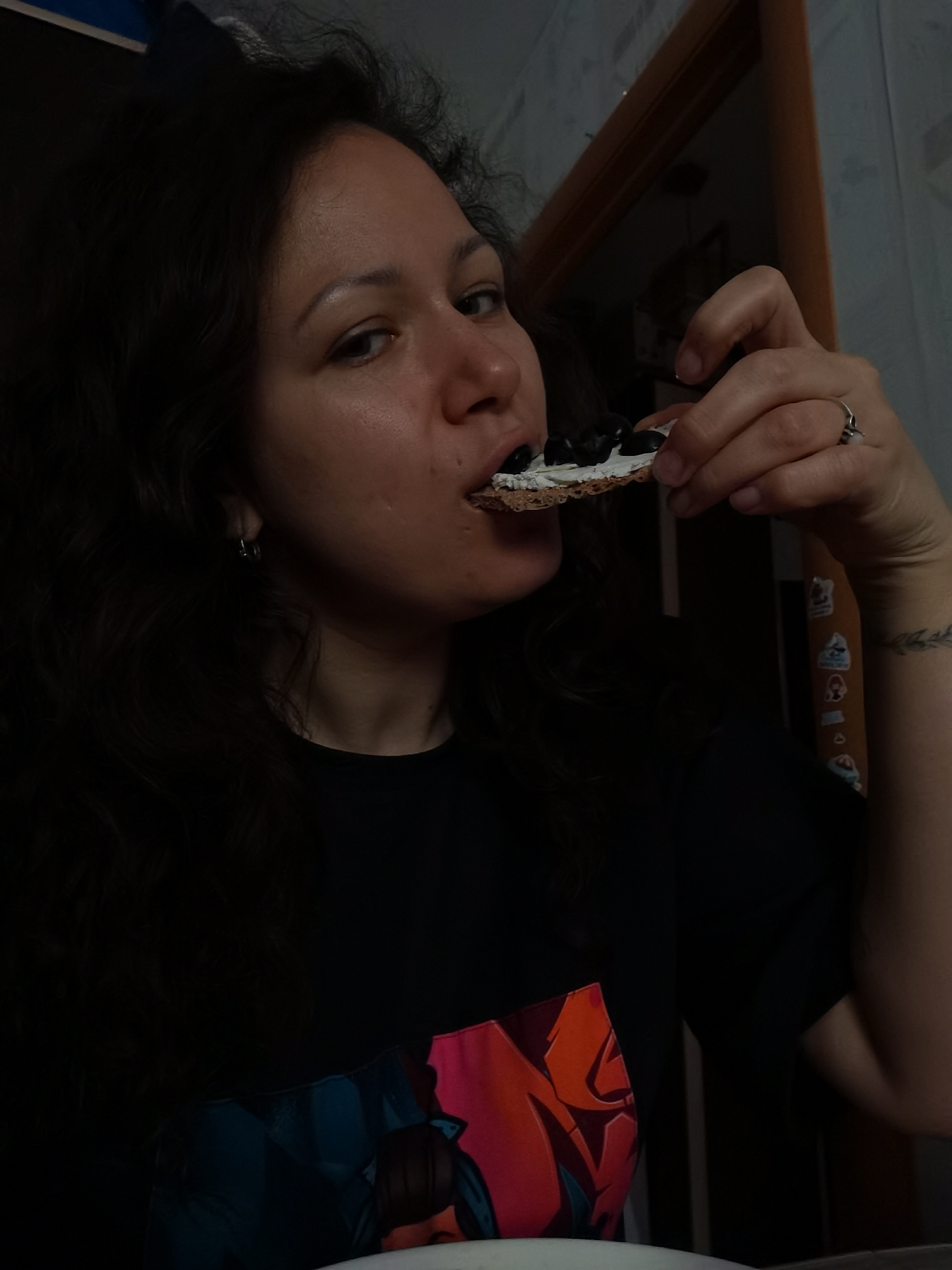 A woman is shown in a close-up shot, holding a cracker topped with a white spread and dark circular objects, likely olives, to her mouth. Her dark, wavy hair frames her face, and she is wearing a black t-shirt with a colorful graphic print on the lower left side. She has a small hoop earring in her left ear and a ring on her right ring finger. A tattoo of a leafy branch is visible on her left forearm. The background is dimly lit, with what appears to be a doorway and wall. The lighting suggests an indoor setting, possibly a kitchen or dining area. The woman's expression is neutral as she is about to eat. There is no discernible information within the image that indicates the location in Chaikovsky, Russia.