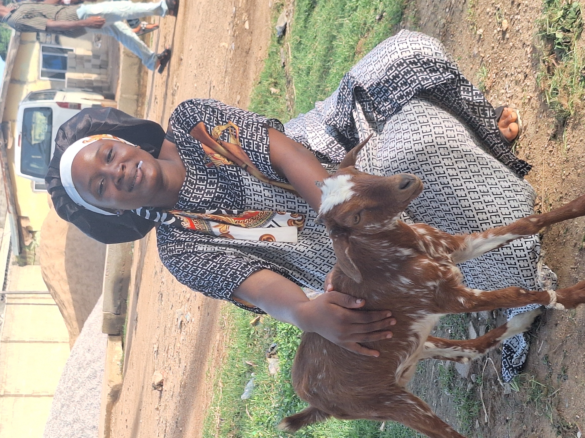 In Sabon Tasha, Nigeria, a woman wearing a black head covering with a white band, and a patterned black and white dress, sits on the ground. She is holding a brown and white goat. A man in jeans walks in the background. A white van is parked near a building. The surrounding environment consists of dirt, grass, and buildings.
