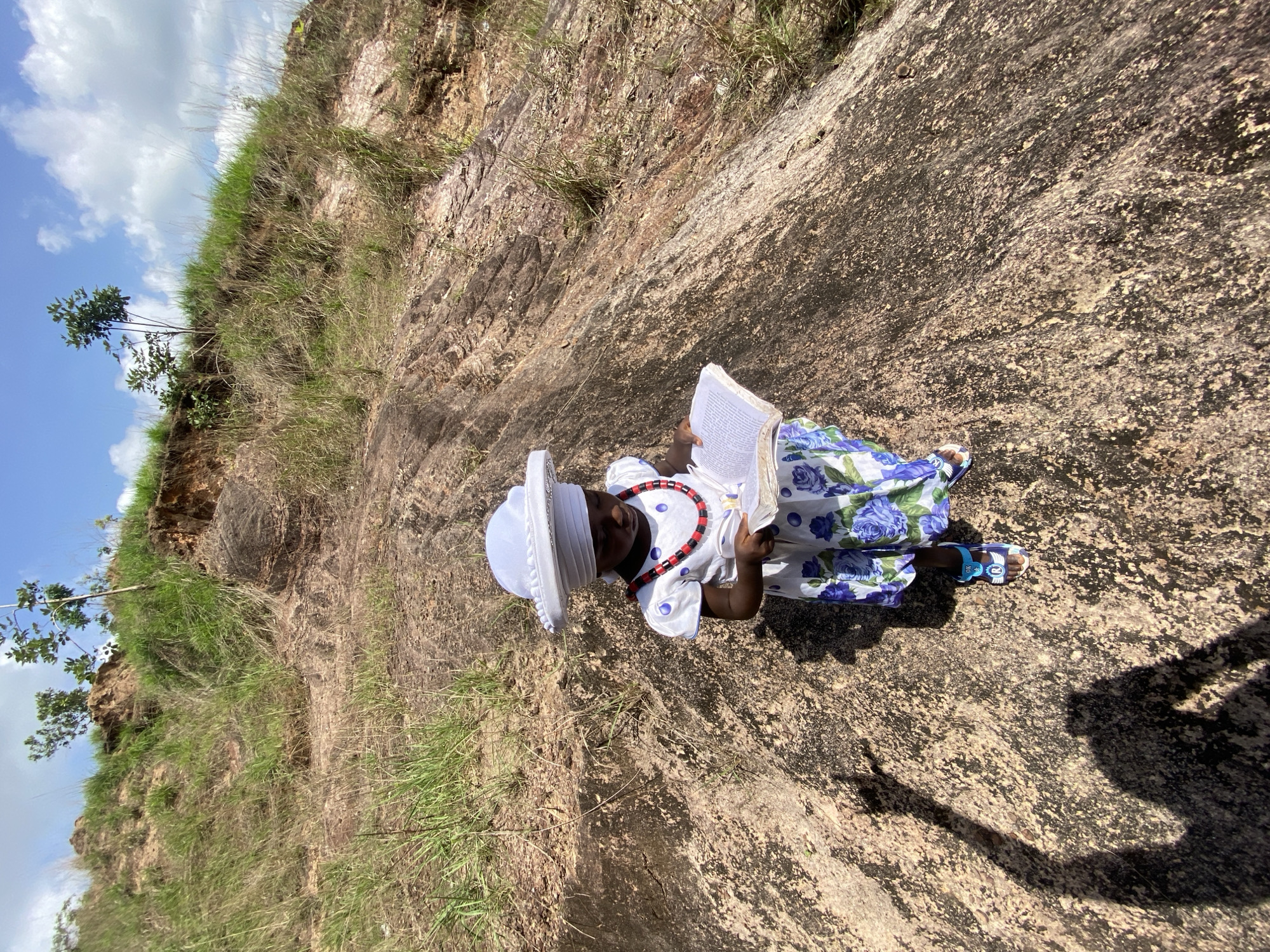 A young girl is sitting on a rocky incline in Amaseri, Nigeria, reading a book. She is wearing a white hat, a patterned dress with blue roses, and sandals. A red and black beaded necklace adorns her neck. The rocky landscape is covered in patches of grass and low-lying vegetation. The sky is visible with some clouds. A shadow is present near her feet.