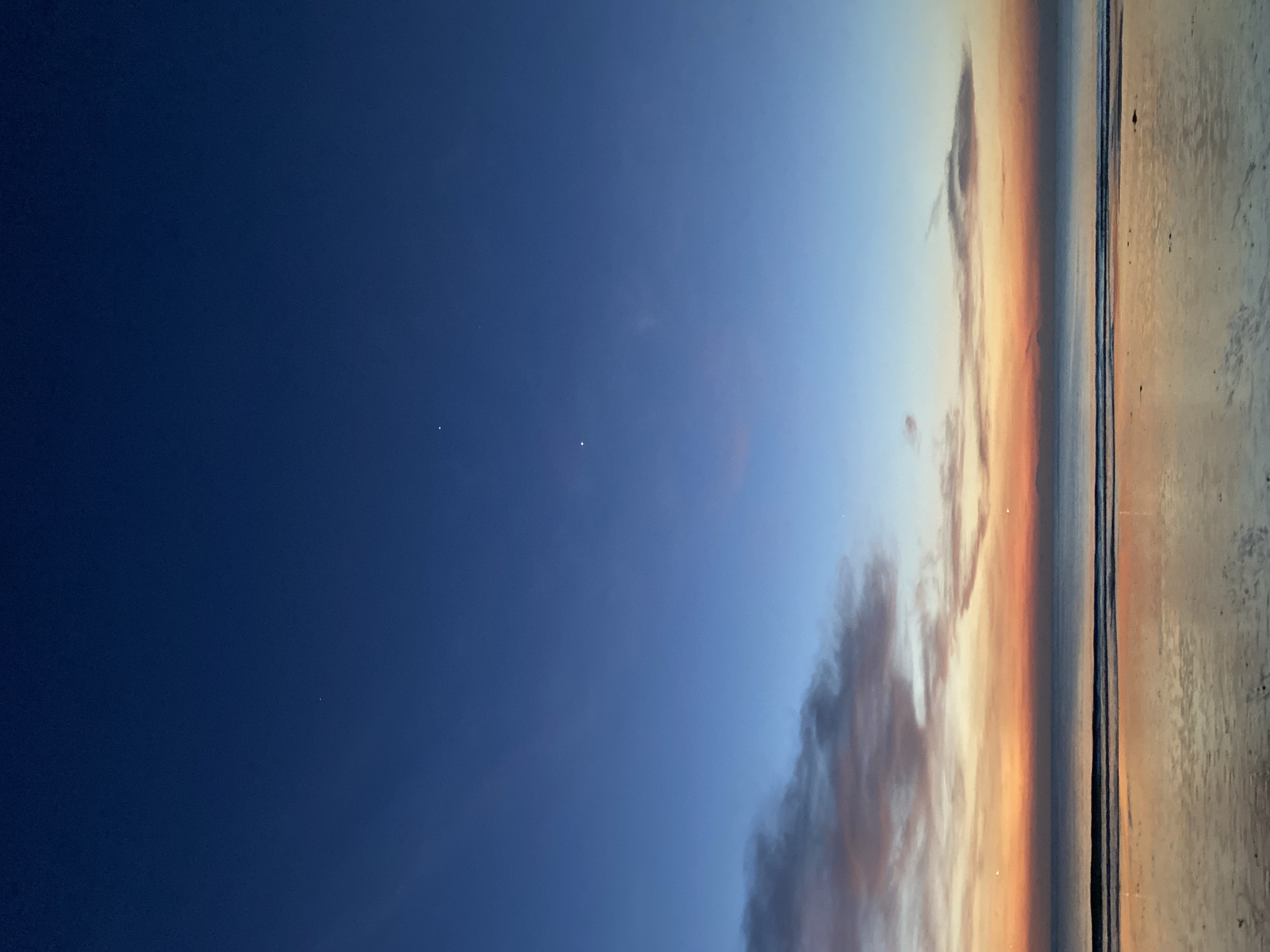 The image depicts a beach scene at dusk in Kuta Selatan, Indonesia. The sky transitions from dark blue to lighter shades with orange hues near the horizon, suggesting a sunset. Wispy clouds are visible in the sky, tinted with orange and brown colors. The coastline is marked by a line of gentle waves meeting the shore. The beach itself appears wet, reflecting the remaining light from the sky. Two small, bright dots are visible in the darker blue portion of the sky. No people or other recognizable objects are visible in the scene.