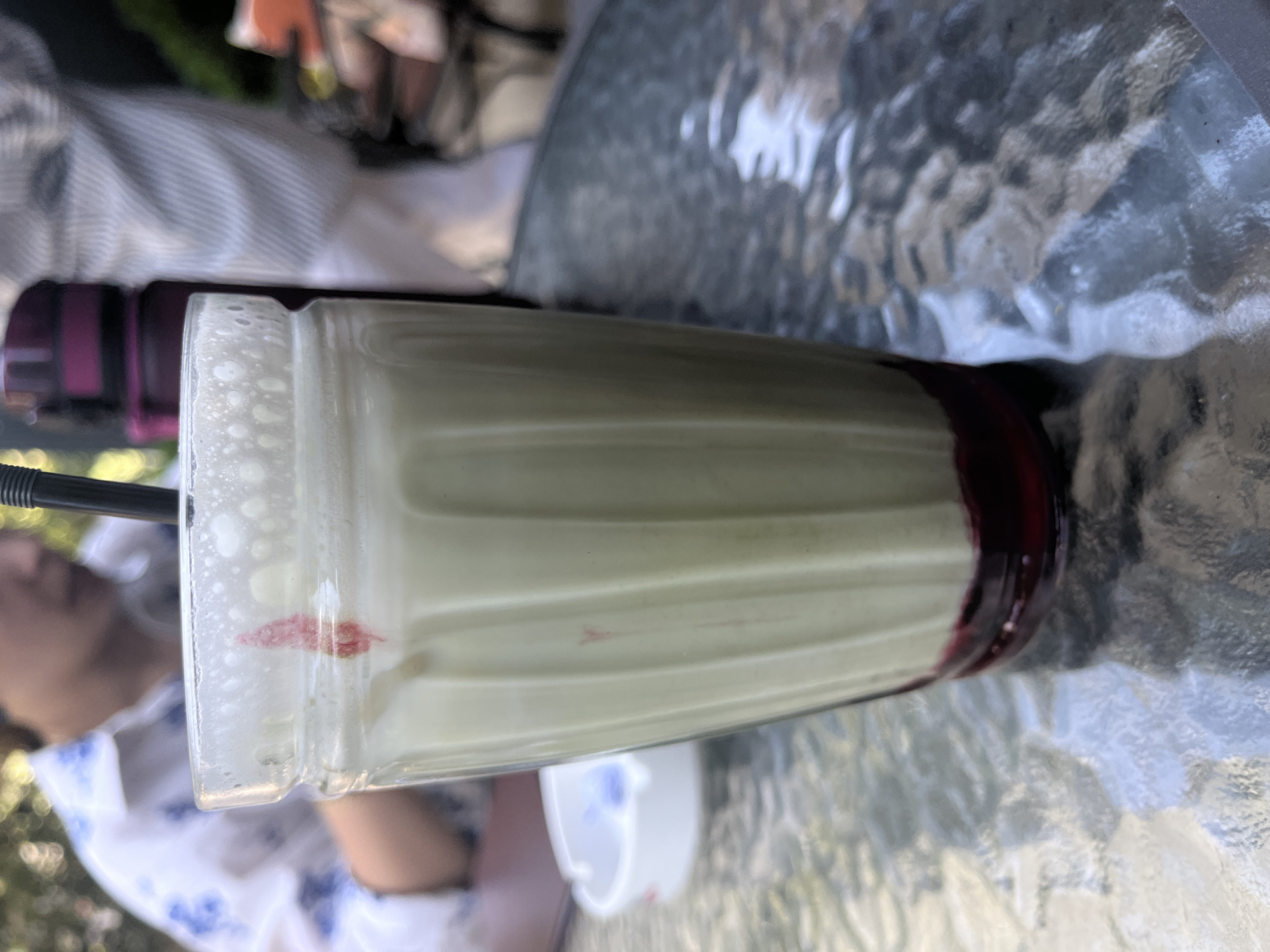 A tall, fluted glass of a pale green beverage with a dark red syrup at the bottom sits on a textured glass tabletop. The glass has a frothy top with a single streak of red on the rim. A black straw is partially visible in the beverage. In the blurred background, a person in a white shirt with blue floral patterns is visible to the left. To the right of the glass, out of focus, are a dark purple thermos and a person wearing a striped shirt. An out-of-focus white ashtray with blue accents is to the left of the glass. The setting appears to be outdoors.