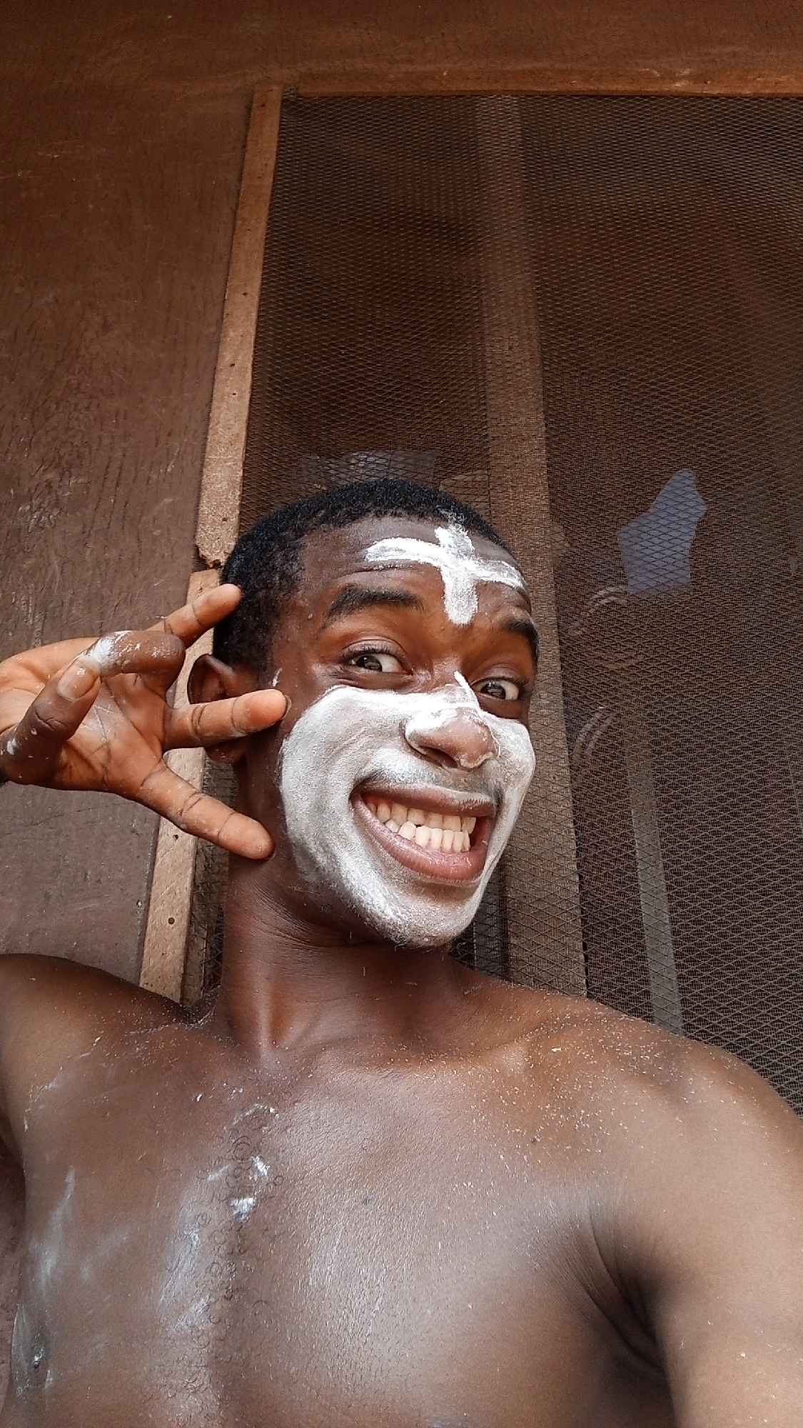 A shirtless, dark-skinned man with a white, mask-like substance on his face and a white cross on his forehead is posing for a selfie. He is smiling widely, revealing his teeth, and holds his left hand near his ear with his fingers extended. The background consists of a screen door and a brown wooden wall. The location is likely inside a building or home, potentially in Onitsha, Nigeria, but there are no distinguishing city features visible in the frame to confirm its location.