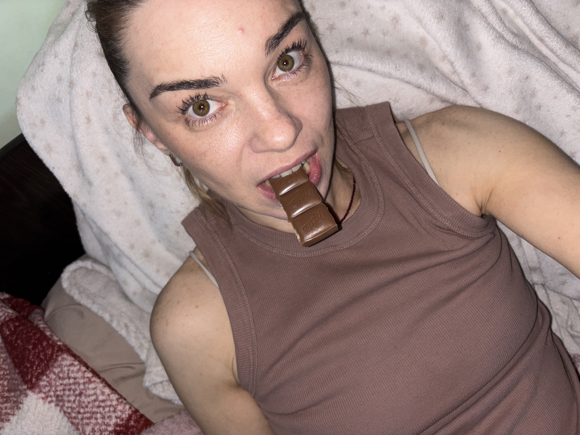 A young adult woman lies on her back on a bed, partially covered by blankets. She is wearing a brown tank top. A segment of a chocolate bar is held in her mouth, between her teeth. Her eyes are open and she is looking towards the camera. The environment appears to be a bedroom, with soft bedding and blankets visible. The lighting is dim and appears to be artificial. The background is dark.
