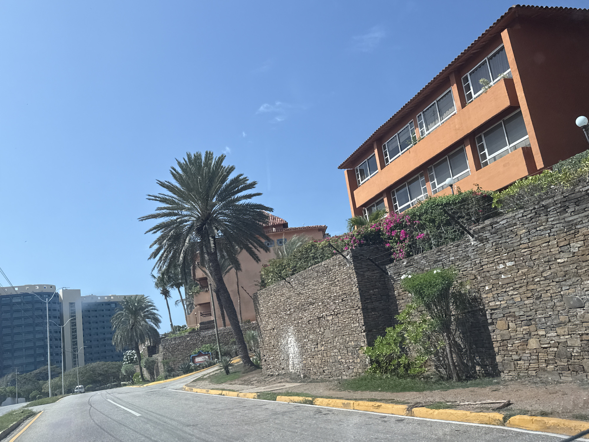The image depicts a street scene in Pampatar, Venezuela, under a clear blue sky. The foreground features a paved road with yellow and white markings, suggesting it's a two-way street. To the right of the road, a dry stone wall runs along an elevated embankment.
On top of the embankment, an orange building with multiple floors and numerous windows is visible, bordered by lush greenery and vibrant pink flowers. A tall palm tree stands prominently in front of the building and the stone wall. In the background, to the left, a cluster of tall, modern apartment buildings with dark facades and a rounded architectural style are visible. Another palm tree is situated between the road and these buildings.
The overall scene suggests a warm, sunny day, likely midday given the bright light and clear skies. The environment appears to be a residential or resort area, characterized by tropical vegetation, modern architecture, and coastal-like stone construction. The presence of the stone wall and the style of the buildings might indicate a location near the coast. No people are visible in the image. The sign on the stone wall, though partially obscured, appears to say "LAS CANAS".
