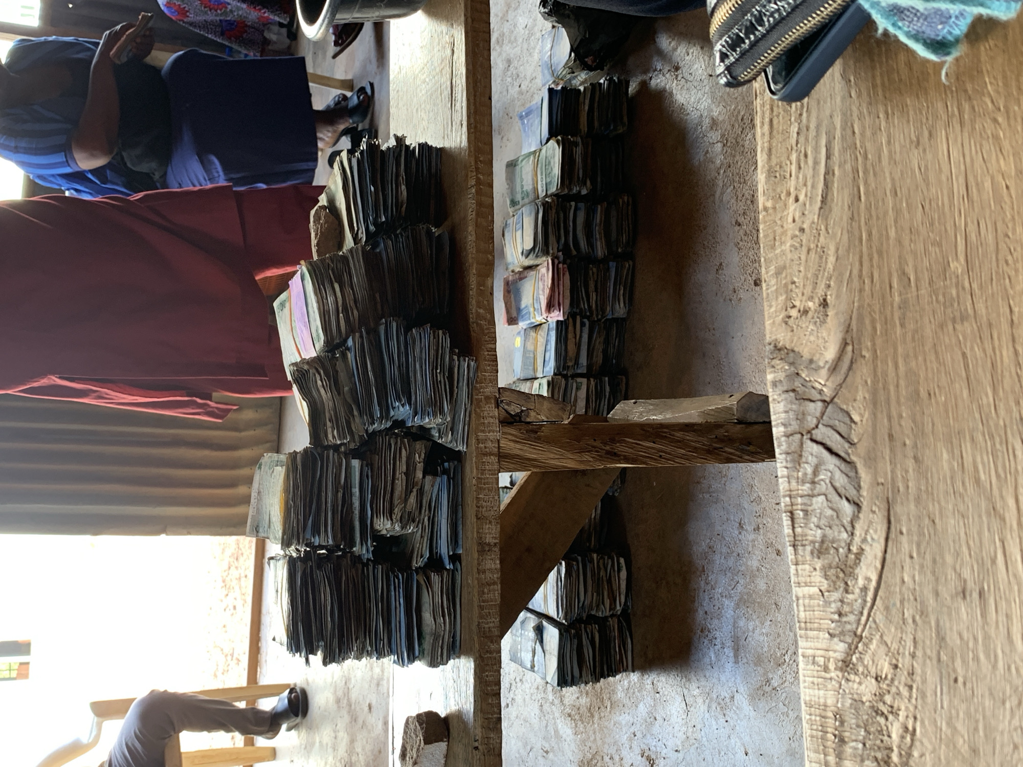 The image shows stacked bundles of currency notes, possibly Nigerian Naira, on a wooden table in Rafin Bauna, Nigeria. A person is seated at the table on the left and another person sitting on another table to the right, with bundles of currency near them. The environment looks to be indoors, possibly a simple building constructed with corrugated metal sheets. The lighting is natural daylight.