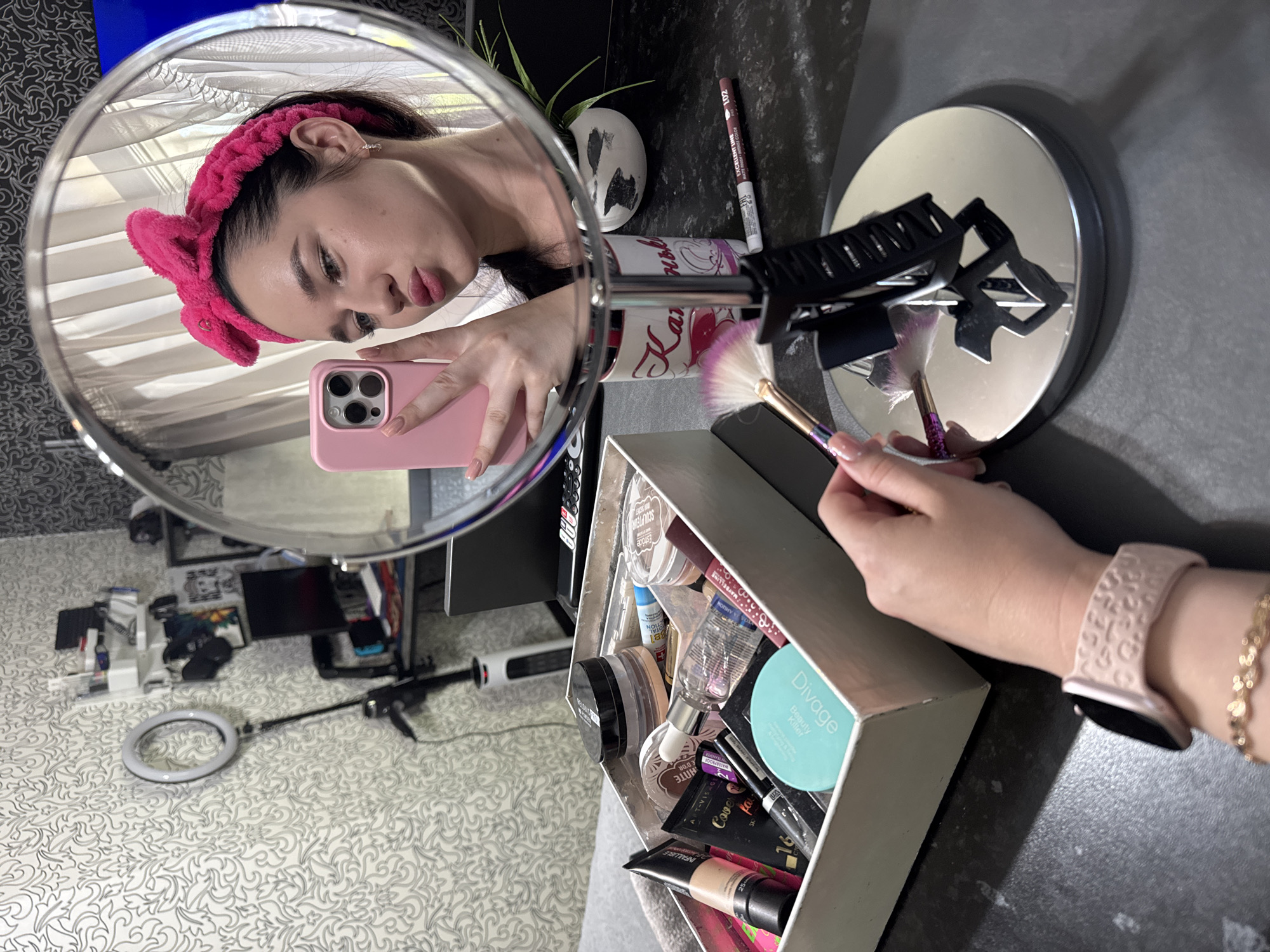 A young woman with a bright pink fuzzy headband, styled in a bow, is taking a selfie in a mirror. She is wearing a dark top and has a pink iPhone in her right hand. Her left hand holds a makeup brush. In the foreground, a makeup box is filled with various cosmetic products. The scene appears to be in a bedroom, with a ring light and shelves with personal items visible in the background, suggesting a personal vanity area.