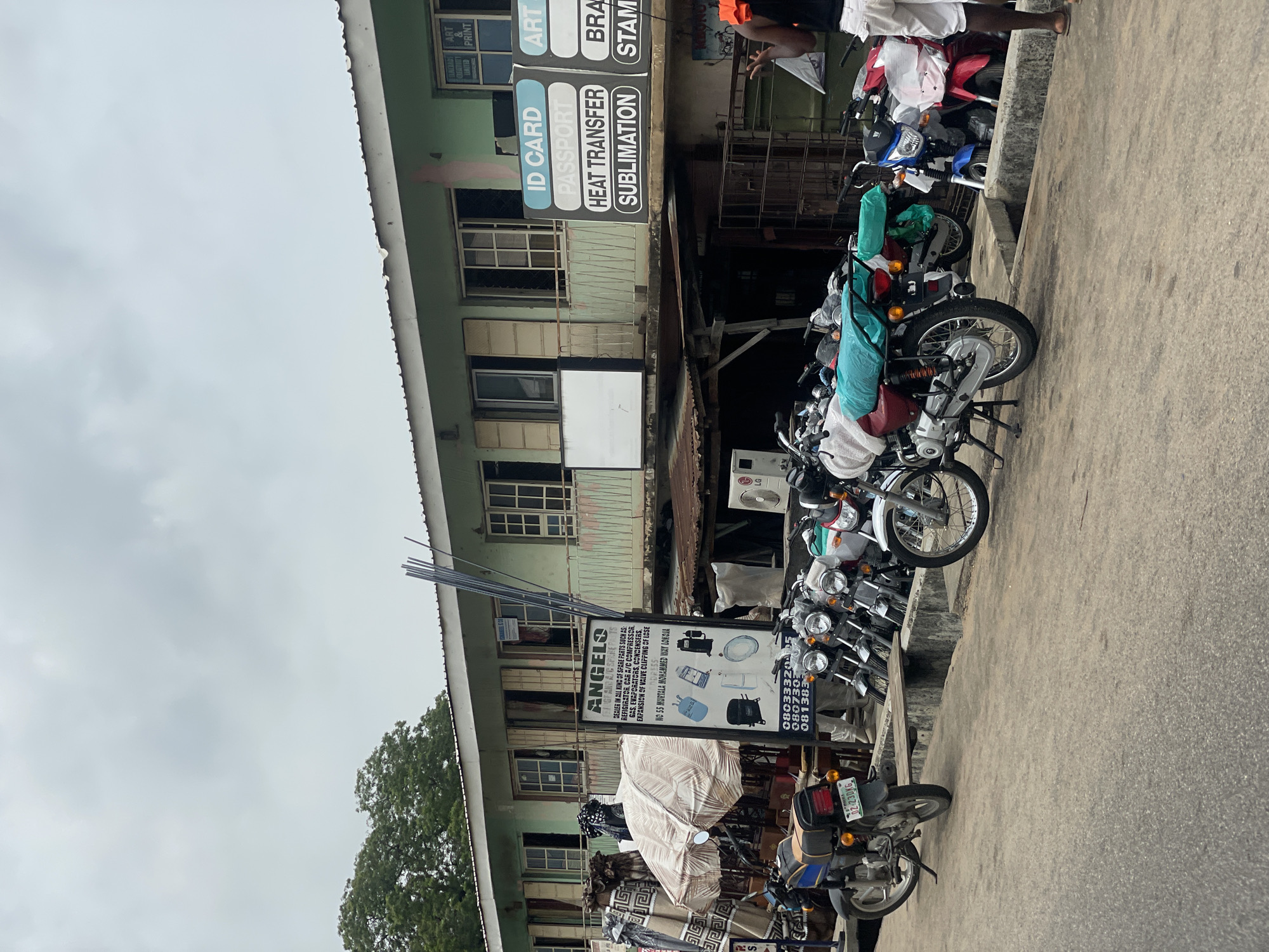 The image captures a street scene in Lokoja, Nigeria, with a focus on a row of parked motorcycles. In the foreground, a paved road leads into the frame, with several motorcycles lined up along a raised concrete barrier. One motorcycle in the foreground is dark blue with yellow and red accents, bearing a license plate that reads "OZ-230 KG NIGERIA". Beyond the motorcycles, a two-story building dominates the background. The building is painted a pale green and has multiple windows, some covered with bars. A sign with the heading "ANGELS" and details about spare parts for air conditioning units is prominently displayed near the center of the image. Another sign to the right advertises "ID CARD", "PASSPORT", "HEAT TRANSFER", and "SUBLIMATION" services, along with "ART & PRINT". The sky is overcast. A few individuals are visible; one person in the foreground on the right is wearing white shorts and a dark top, and appears to be gesturing with their right hand.