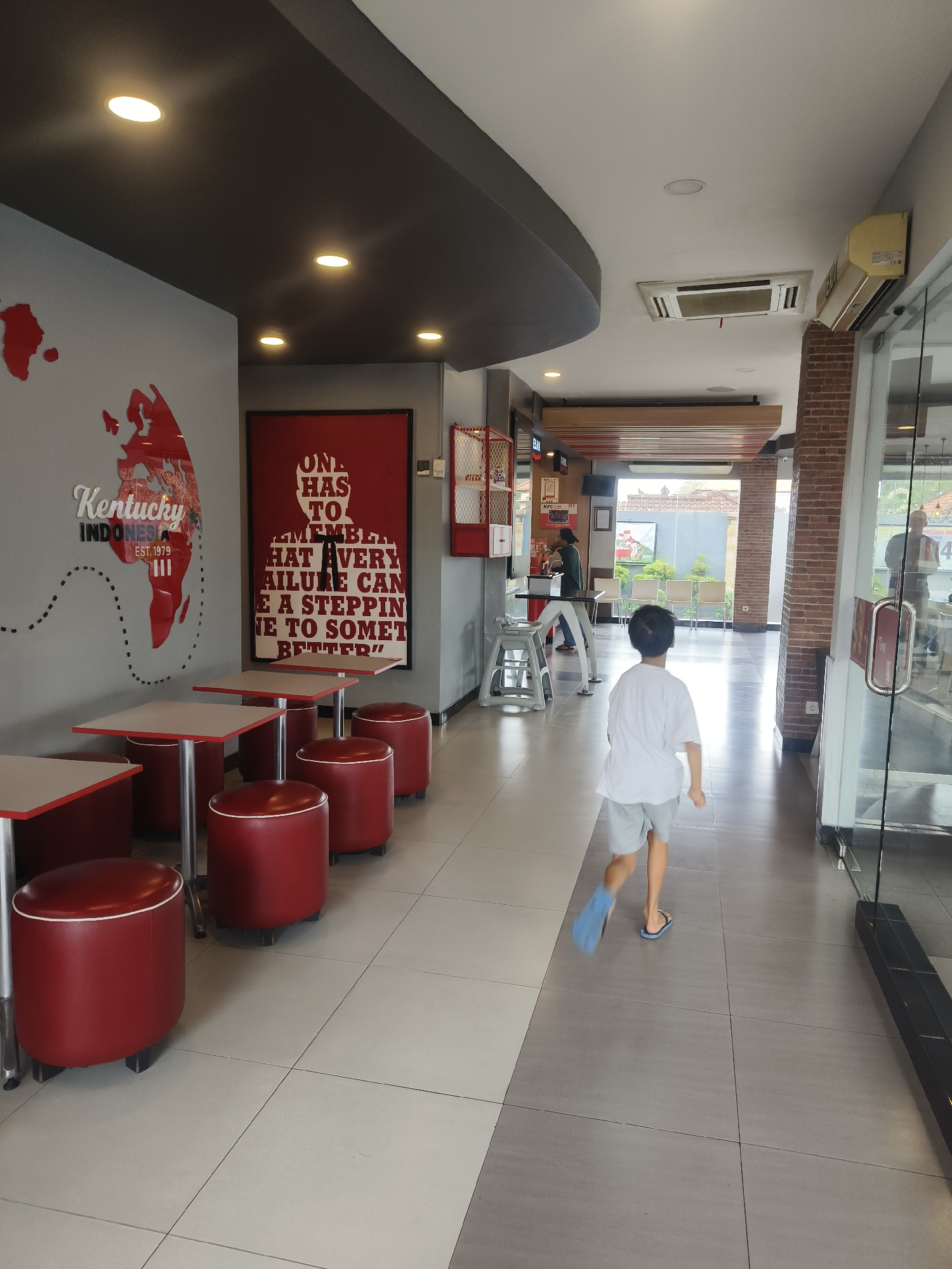 The image captures the interior of a KFC restaurant in Denpasar, Indonesia. A young boy, wearing a white t-shirt and grey shorts, is seen running away from the camera down a tiled hallway. He is wearing blue flip-flops. The hallway is flanked by tables with red stools and square tabletops on the left, and a brick-patterned wall and glass doors on the right. A world map graphic with "Kentucky Indonesia est. 1979" is visible on the left wall. A large framed quote is also on the left wall. In the background, other customers and staff are visible, and natural light streams in from an open doorway. The ceiling features recessed lighting and an air conditioning unit.