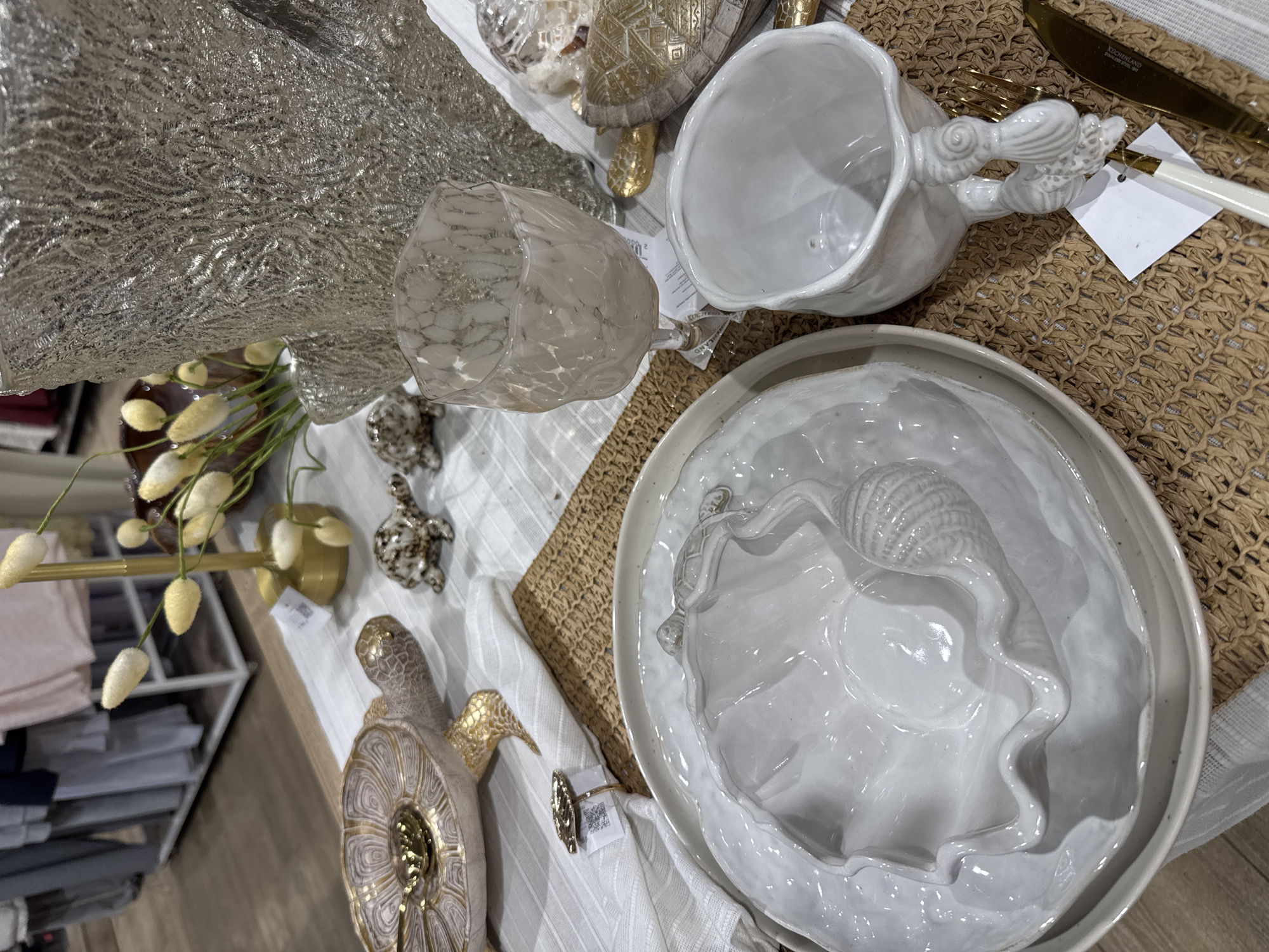 The image shows a table setting, likely indoors, featuring various decorative objects. There's a silver textured material, possibly fabric or a decorative item, and a small vase holding faux floral stems with white buds. Additionally, there are two decorative turtle figurines, one with gold accents, and a marbled glass.
The place setting includes a white, wavy-edged plate with a seashell design in the center, a white mug with a bird-shaped handle, and gold-colored cutlery. A small square tag is attached to the cutlery. The objects are arranged on a table covered with a textured mat and a white cloth with subtle stripes. A shelving unit filled with folded linens is partially visible in the background, suggesting this could be a retail display.
The image does not provide sufficient information to determine the specific location, including if it is in Saint Petersburg, Russia.