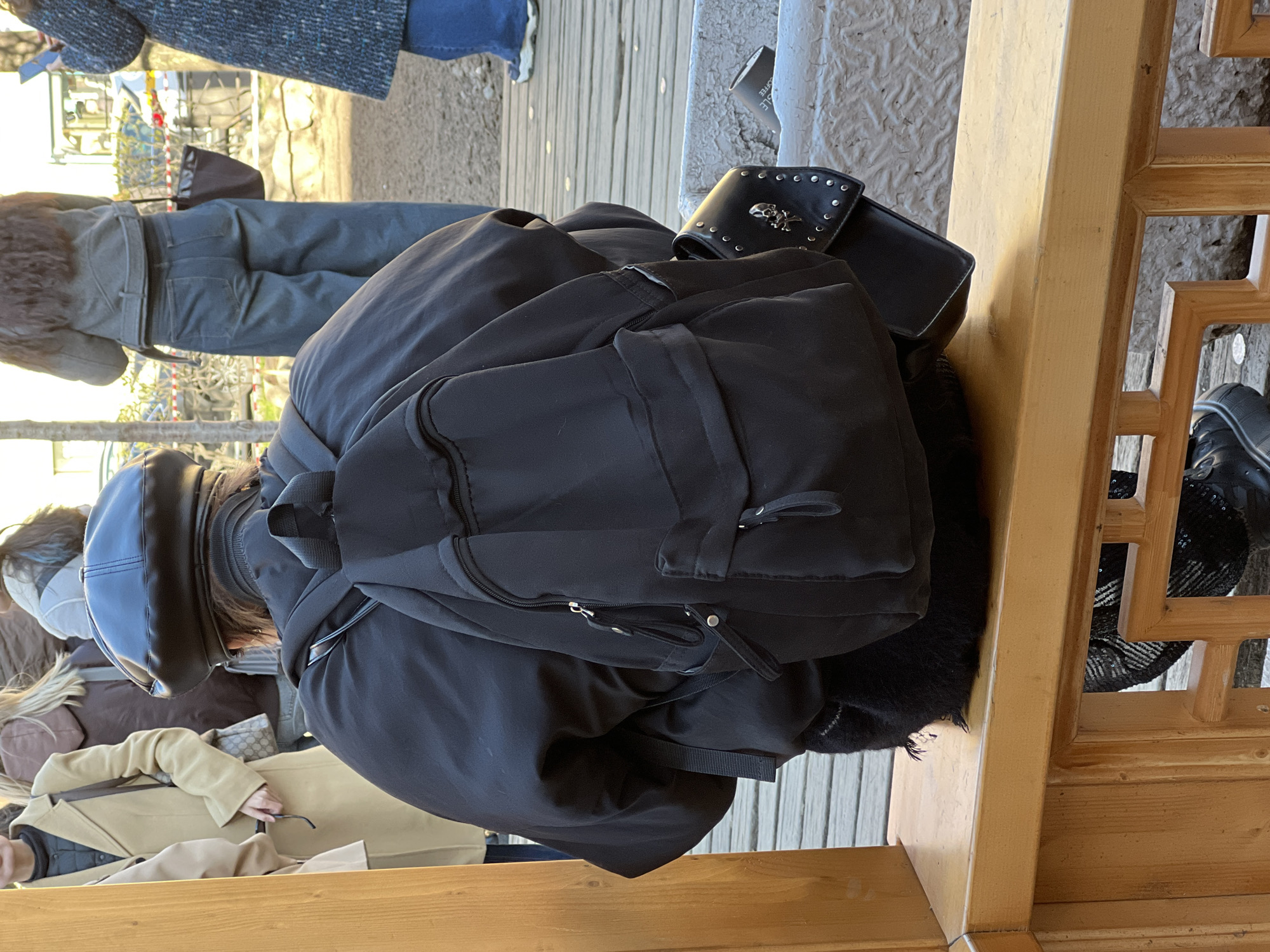 The image captures an outdoor scene in what appears to be a public area in Saint Petersburg, Russia, under bright daylight. The main subject is a person viewed from behind, wearing a black puffer jacket, a black backpack, and a black beret-style hat. Their brown hair is visible beneath the hat.
The person appears to be sitting or leaning against a light-colored wooden structure, possibly a bench or a decorative barrier, which features intricate lattice patterns. Next to the main subject, on the wooden structure, rests a black studded wallet or clutch bag adorned with a silver skull motif. A coffee cup with the word "COFFEE" visible on its side is also present nearby.
In the background and surrounding areas, several other individuals are visible. One person is seen from the back, wearing dark trousers or jeans and a greyish-brown jacket. Further to the left, another person wears a beige trench coat, and fragments of other people and their clothing, including blue jeans and a textured grey coat, can be seen. The ground appears to be a mix of light-colored paving or cobblestones and darker wooden planks. The clothing worn by the individuals suggests cool weather. The overall setting implies a busy outdoor space, possibly a park or tourist area, with people engaged in casual activities like walking or resting.