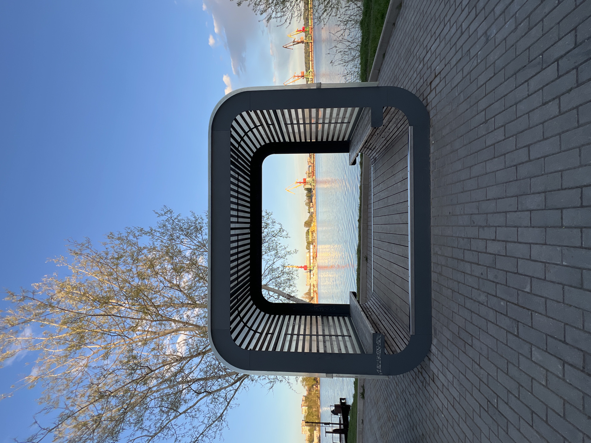 The image depicts a modern, cube-shaped bench structure positioned on a brick-paved path. The bench has a gray frame with horizontal slats and a wooden seat. Through the structure, there is a view of a river with cranes visible in the distance. A tree with bare branches is located to the left of the bench. The sky is blue with some scattered clouds. The environment suggests an urban waterfront location. According to geolocation data, it is located at a modern park called "Sovietsky" in Cherepovets, Russia.