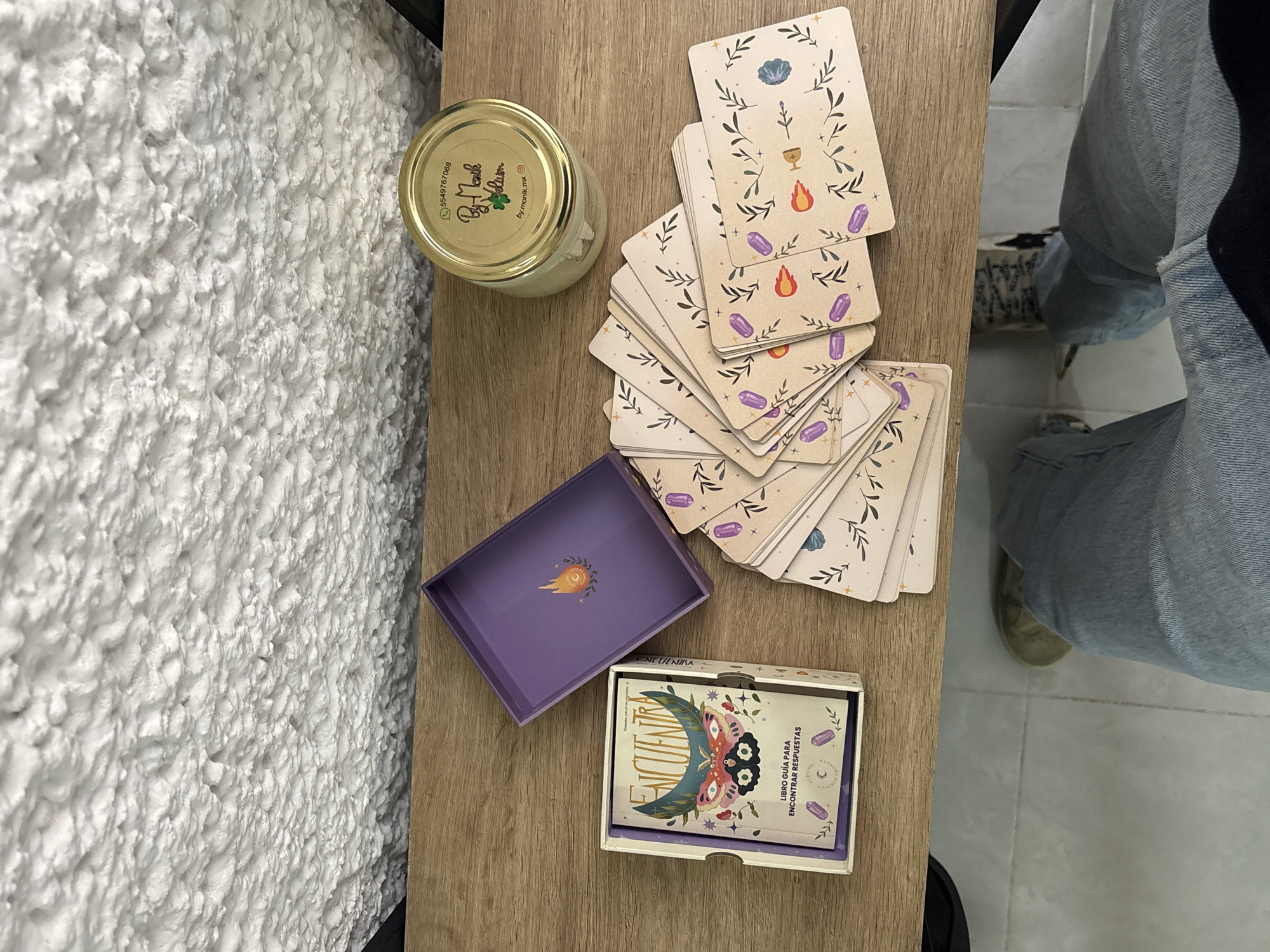 An overhead indoor shot captures a collection of items arranged on a light-colored wooden surface, next to a highly textured white wall. On the left, a glass jar with a golden lid is visible. The lid displays the text "5549767068", "By Monk", "by.monik.mx", along with an Instagram icon and a small plant illustration.
To the right of the jar, a deck of elegantly designed cards is spread across the wooden surface. These cards are adorned with botanical motifs, including leaves and branches, and feature various symbols such as a shell, a chalice, a sword, flames, and purple crystals. Below the main card arrangement, a vibrant purple box, likely part of the card set, is open, revealing a stylized comet or shooting star design on its interior lid.
Next to the open purple box, a smaller white box holds a guidebook. The guidebook's cover showcases a detailed moth illustration set against a crescent moon, with the prominent title "ENCUENTRA". Additional Spanish text on the guidebook reads "LIBRO GUÍA PARA ENCONTRAR RESPUESTAS" (Guidebook to Find Answers) and "ALHUMO".
In the lower right portion of the image, the legs of a person wearing blue jeans and casual shoes are partially visible, indicating a presence nearby, perhaps seated or standing. The overall lighting suggests a daytime setting, though no specific time or outdoor weather conditions are discernible. The Spanish text on the card set components is consistent with the provided location context of Puebla, Mexico. The arrangement of the cards and guidebook suggests an activity involving introspection, guidance, or a game.