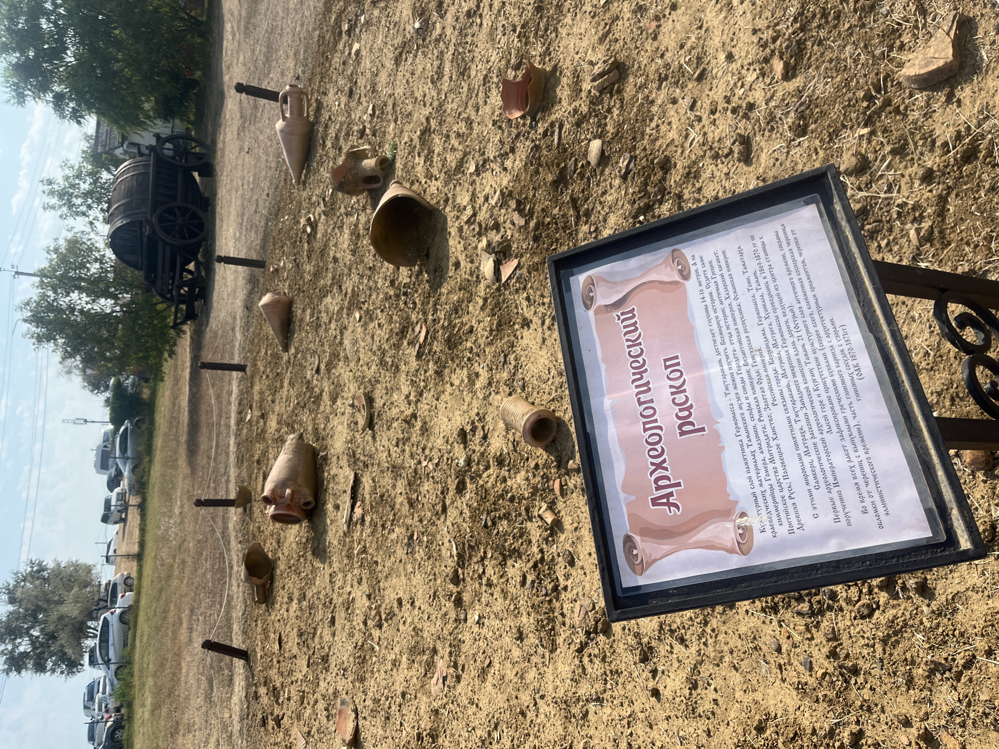 An outdoor scene in Taman', Russia features an archaeological dig site with numerous broken clay amphorae and pottery shards scattered across sandy soil. In the foreground, a dark-framed informational sign with Cyrillic text stands upright. The sign has a scroll-like graphic and the title "Археологический раскоп" (Archaeological excavation). The background shows a dry, grassy embankment with several parked cars, trees, and a rustic wooden wagon with large wheels. The sky is visible above, with scattered clouds.