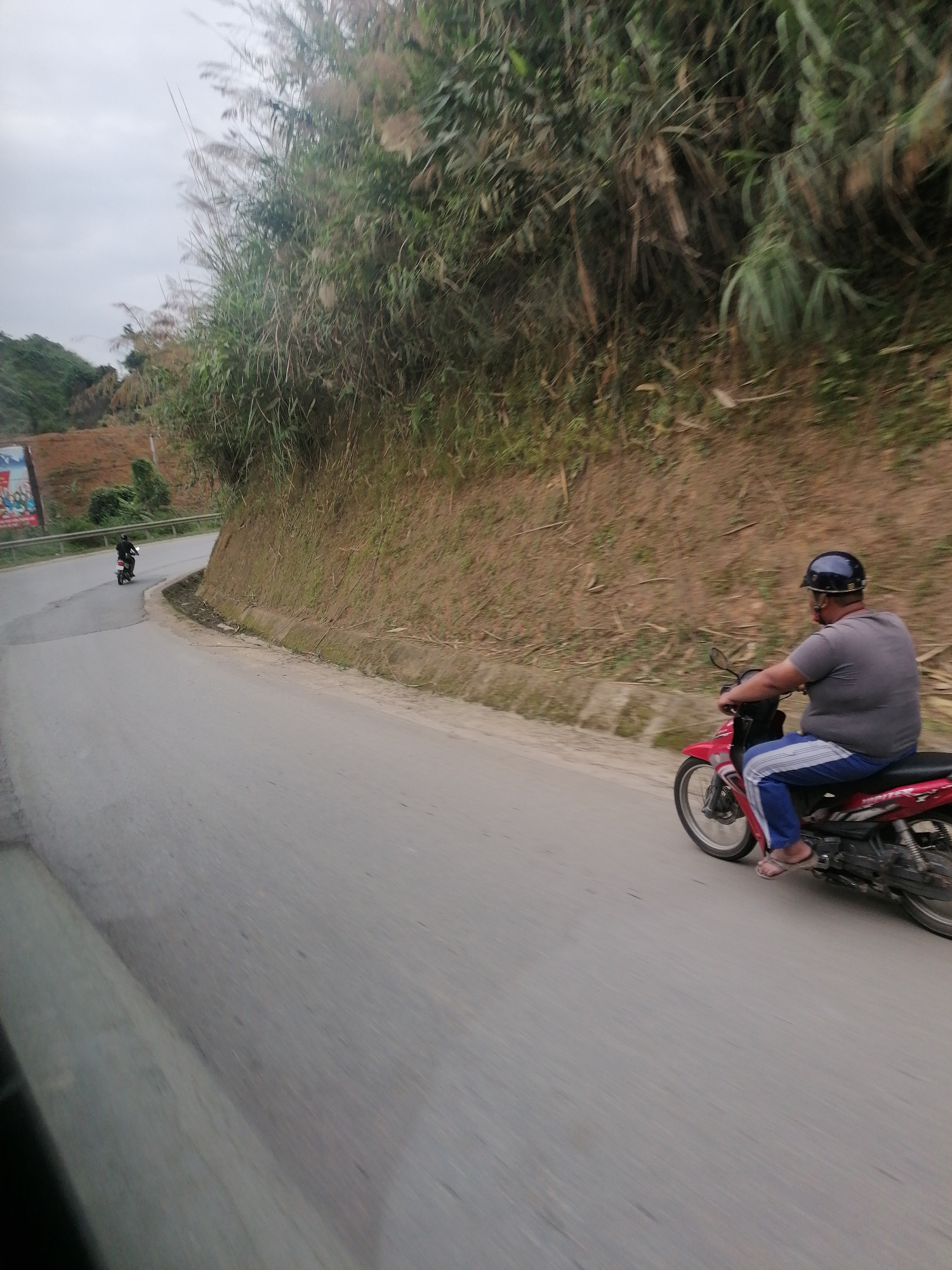 The image captures a street scene in Cốc San, Vietnam. The road curves around a hillside covered in dense vegetation, with a small billboard partially visible in the background. Two individuals are riding motorcycles. One is closer to the camera, wearing a gray shirt, blue pants with a white stripe, flip-flops, and a dark helmet. The other, further down the road, is wearing all black. The road is paved and appears to be in decent condition. The sky is overcast. The perspective suggests the image was taken from a moving vehicle.