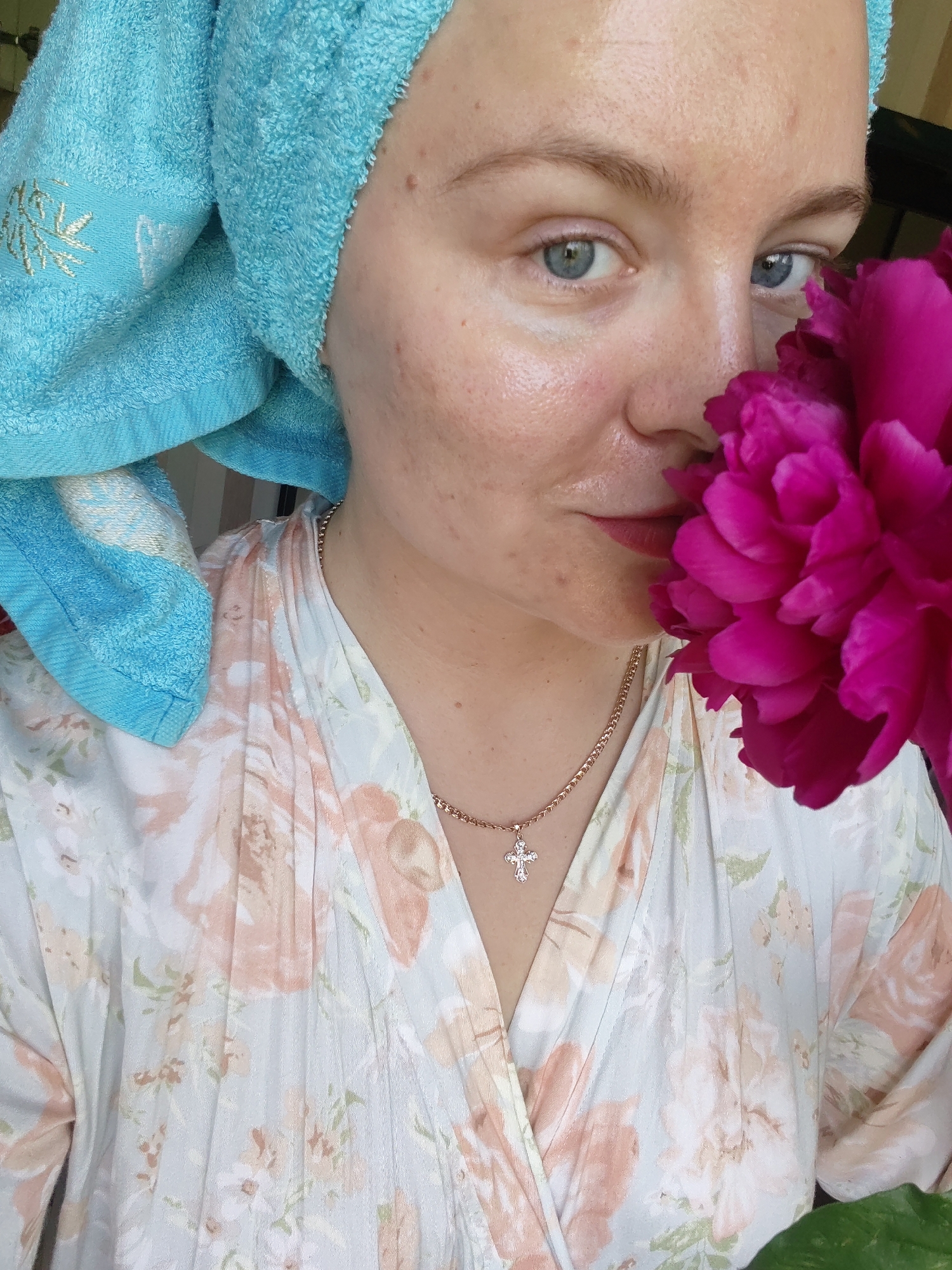 A woman with blue eyes and light skin, wearing a blue towel on her head and a floral robe, is holding a large pink flower up to her face. She is wearing a gold necklace with a cross pendant. The image appears to be taken indoors, possibly in a home in Izhevsk, Russia.