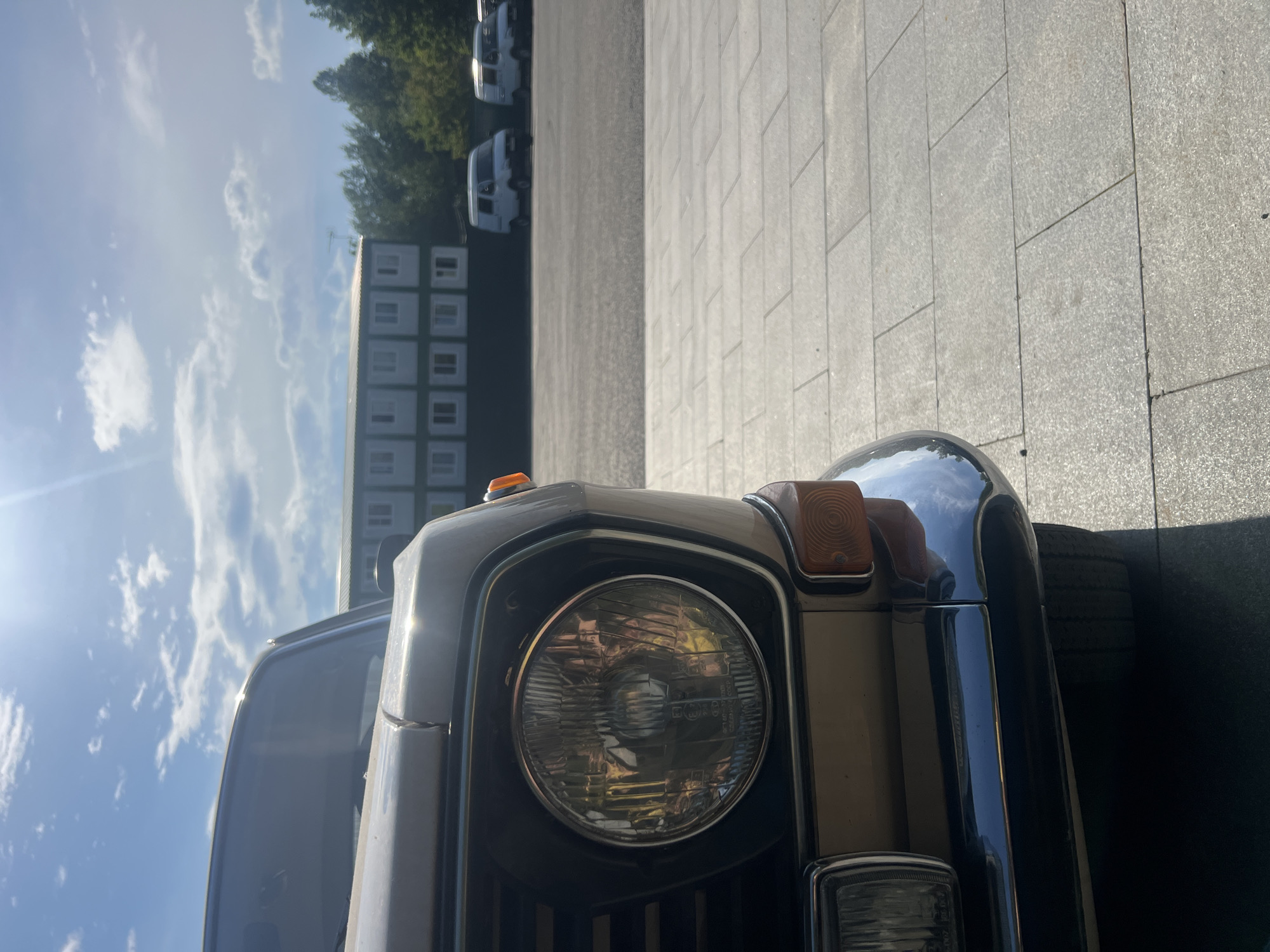 The image shows the front of a classic car, likely a Lada, parked on a paved area. The car is a light brown or beige color with a chrome bumper and a round headlight. An orange turn signal is visible above the headlight. In the background, there are several white modular buildings arranged in rows, possibly temporary housing or storage units. Beyond the buildings, there are trees and a bright blue sky with scattered white clouds, suggesting it is a sunny day. Two white vans are parked in the distance. The sun is low in the sky, casting long shadows and creating a lens flare on the left side of the image, indicating it is either early morning or late afternoon. The scene appears to be an outdoor parking lot or service area. There is no visible text in the image.