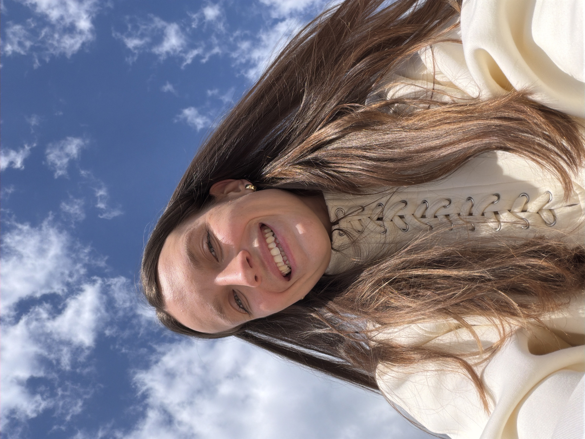 A low-angle view of a smiling woman against a blue sky with white clouds. Her brown hair frames her face, and she is wearing a white top with a laced front. The location appears to be outdoors, possibly Drobnići, Montenegro, given the prompt's instruction. The sky and clouds suggest fair weather. The focus is on the woman's face and the sky behind her.