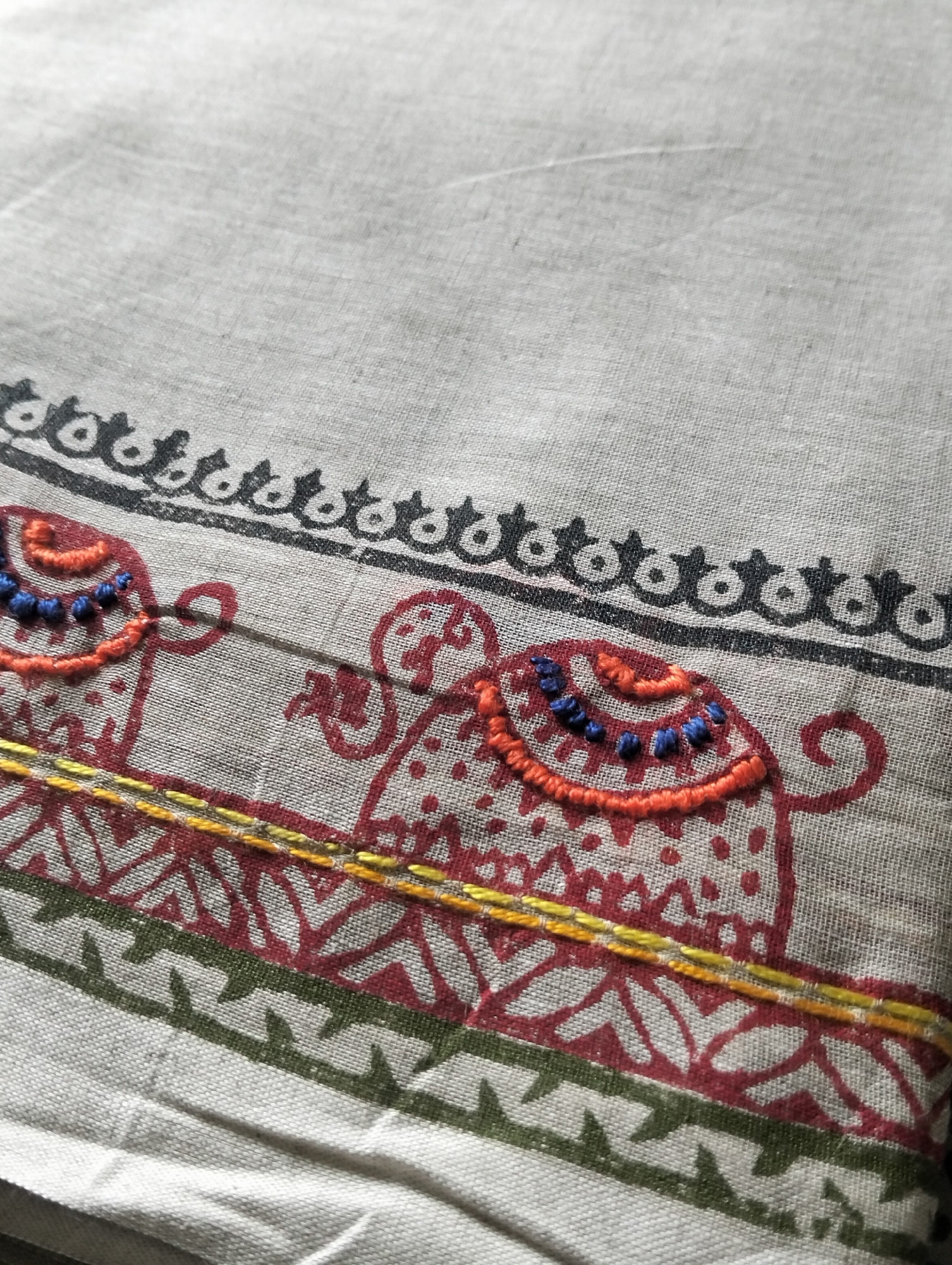 The image shows a piece of handloom fabric, likely cotton or linen, with a block-printed design featuring elephants. The print colors include red, green, and black. The elephants' headgear is decorated with orange embroidery and blue beads. A yellow embroidered line separates the elephant pattern from a lower geometric pattern. The location in Dhaka, Bangladesh is inferred from the style of block printing and embroidery, which is a common craft in that region.