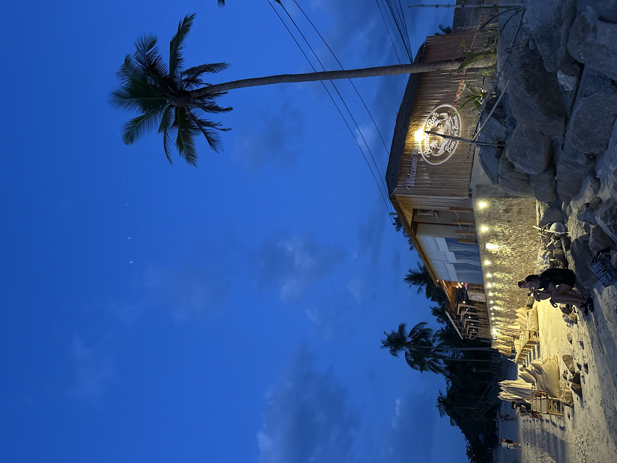 In the city of Ko Samui District, Thailand, a beachfront bar named "Bamboo Bar, Bang Por Beach" is illuminated by string lights and a spotlight under a twilight sky. The bar, constructed with bamboo walls and a thatched roof, features a circular logo with white text. In front of the bar, loungers and beach umbrellas are set up on a sandy beach. Two individuals are seated on rocks in the foreground, facing the bar, while other people are visible in the distance, walking along the shore. A tall palm tree stands prominently beside the bar, with more palm trees forming a silhouette against the darkening sky, which is dotted with faint clouds and a crescent moon. Electrical wires are visible running across the scene.