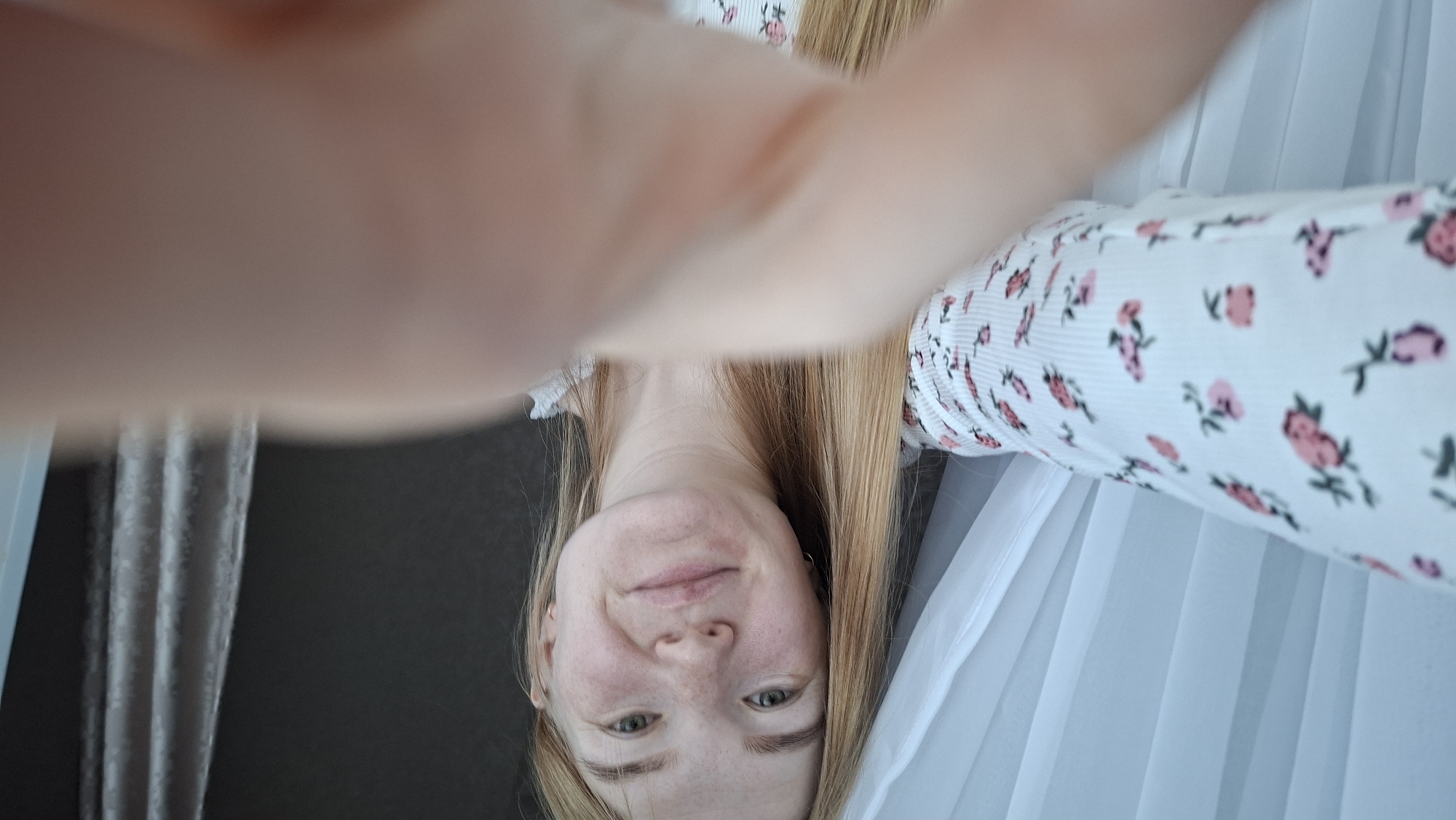 An indoor, close-up, first-person perspective shot captures a young adult female looking directly at the camera. She has long, straight, light blonde or light brown hair and fair skin with visible freckles across her nose and cheeks. Her eyes are light-colored, appearing green or hazel. She has a neutral expression with a slight, subtle smile and is wearing small, gold-colored hoop earrings in both ears. Her attire consists of a white, ribbed top or blouse patterned with small, pink and reddish-pink floral motifs, depicting roses and leaves.
The foreground is partially obscured by an out-of-focus hand, likely belonging to the subject, holding the camera or phone. The background to the left features white, translucent, possibly sheer curtains, suggesting a window and source of soft natural light. To the right, the background is a dark, solid surface, likely a wall, with a segment of a textured, possibly heavier, light-colored curtain visible further to the right. The visual elements within the image do not provide any specific indicators to identify the location as the city of Chaikovsky, Russia.