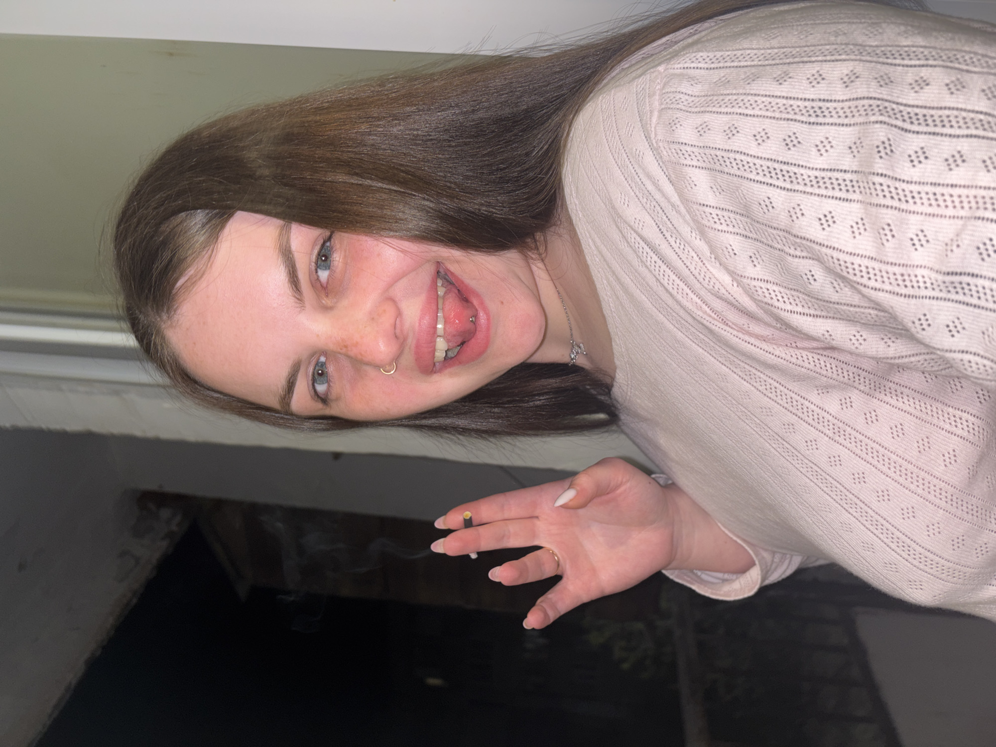 The image, rotated 90 degrees clockwise, presents a young woman smiling broadly at the camera. She has long, dark brown hair, blue eyes, and visible freckles across her nose and cheeks. Her mouth is open in a wide smile, revealing her teeth and a silver barbell piercing on her tongue. She also has a gold hoop piercing in her right nostril.
She is wearing a light-colored, possibly pale pink or beige, long-sleeved top with a distinctive open-knit or lace pattern. A delicate silver necklace is visible around her neck. In her right hand, held casually with white-painted fingernails, she holds a lit cigarette or joint, with a wisp of smoke rising from it. A simple ring is visible on her right ring finger.
The background is dark and indistinct, suggesting an outdoor, nighttime setting, possibly near a balcony or window, with a light-colored structure above her head. The woman appears happy and relaxed. There is no visible text. The activity depicted is a casual moment of smoking, likely taken during the night. The location context for this image is Chornomorskoe, Ukraine.