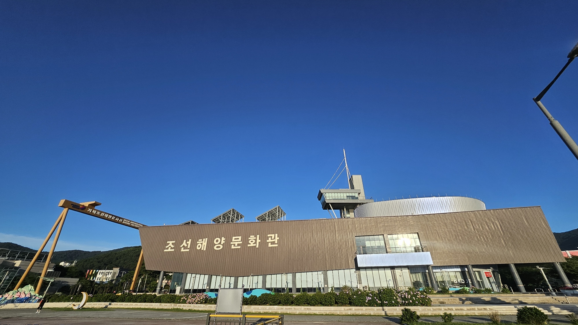 The image shows a wide, low-angle shot of the Geoje Marine Culture Museum in Geoje-si, South Korea, under a clear blue sky. The museum is a modern building with a prominent, slightly curved, brown facade featuring large windows and a metallic roof structure with solar panels. To the left of the main building, a large, yellow, arch-like structure with text on it stands, possibly an entrance or art installation. In front of the museum, there are manicured bushes and flowers, and a paved walkway. A street lamp is visible in the upper right corner of the frame. In the background, hills covered in trees are visible. A lone person is visible on the walkway in the lower left quadrant of the image.